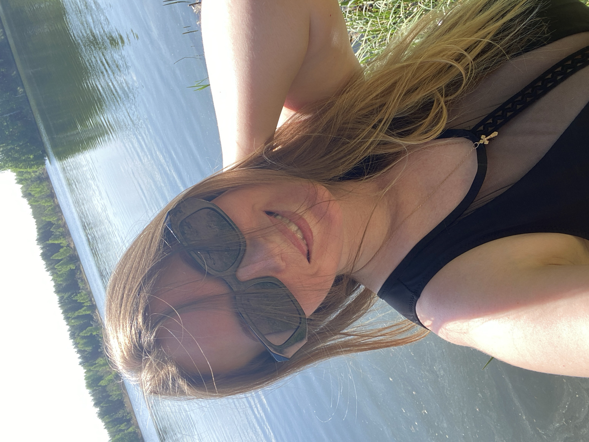 A woman wearing sunglasses and a black swimsuit stands near a body of water. The background reveals a forest and sky, suggesting the scene is likely outdoors in a natural environment. Based on the prompt information, this location is in Novyi, Russia.