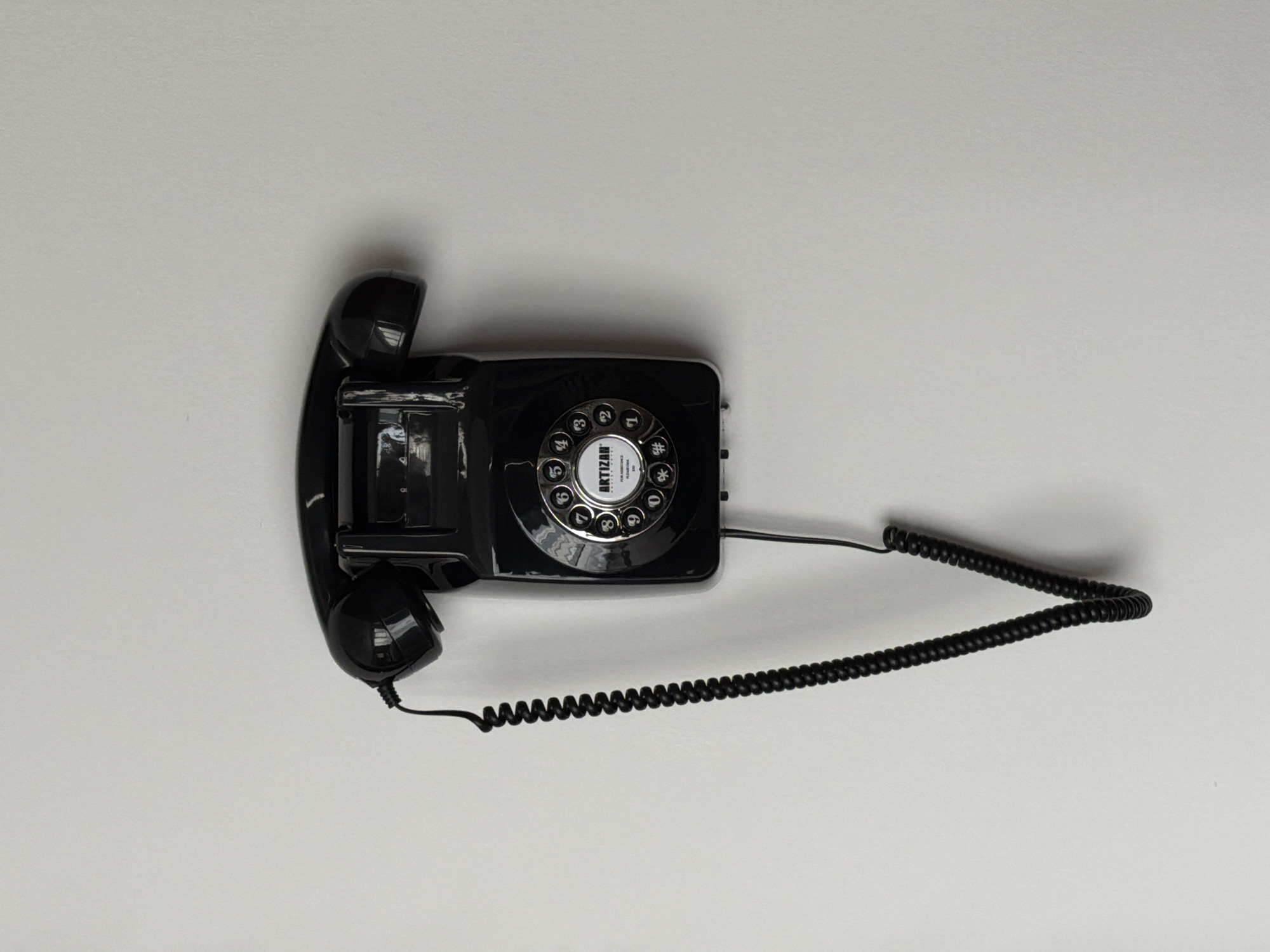 A black rotary dial telephone is mounted on a white wall. The telephone has a coiled cord that hangs down to the left. The dial features numbers from 0 to 9, as well as symbols for asterisk and pound. The word "ARTIZAN" is visible in the center of the dial. The background is a plain white wall. There are no people or other objects visible in the image. The location is not specified as being in Tbilisi, Georgia.