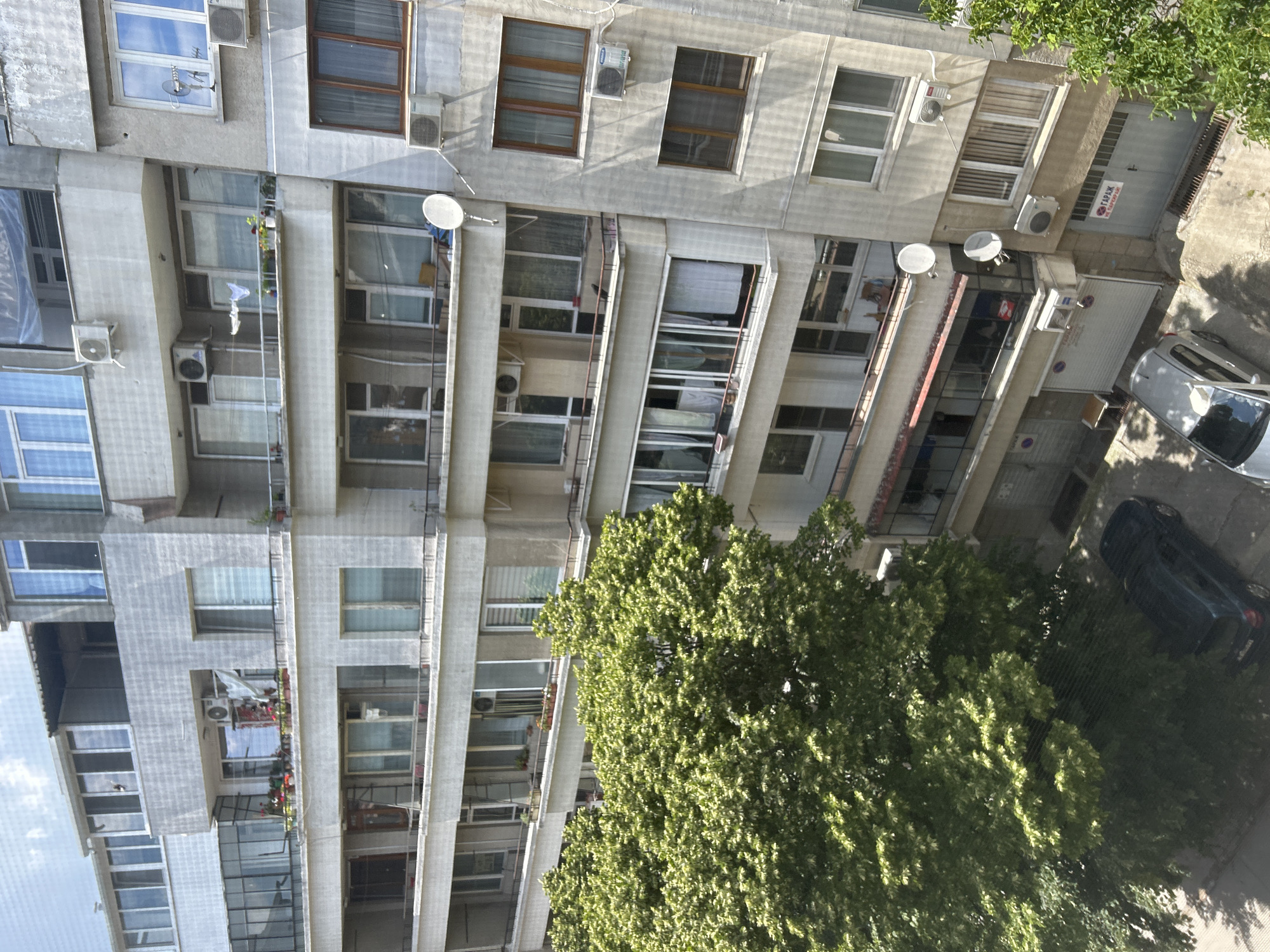 The image captures a view of an apartment building in the city of Varna, Bulgaria. The building appears to be several stories tall, with multiple balconies and windows visible. Some balconies are adorned with potted plants, adding a touch of greenery to the otherwise concrete structure. Air conditioning units are mounted on the exterior walls of several apartments.
In the foreground, a large, leafy green tree partially obscures the lower floors of the building. Below the tree, on the street level, two cars are parked. One is a dark-colored sedan, and the other is a lighter-colored hatchback. Garage doors are visible on the ground floor of the building, with one bearing a red and white sign. The sky above is partially clouded.