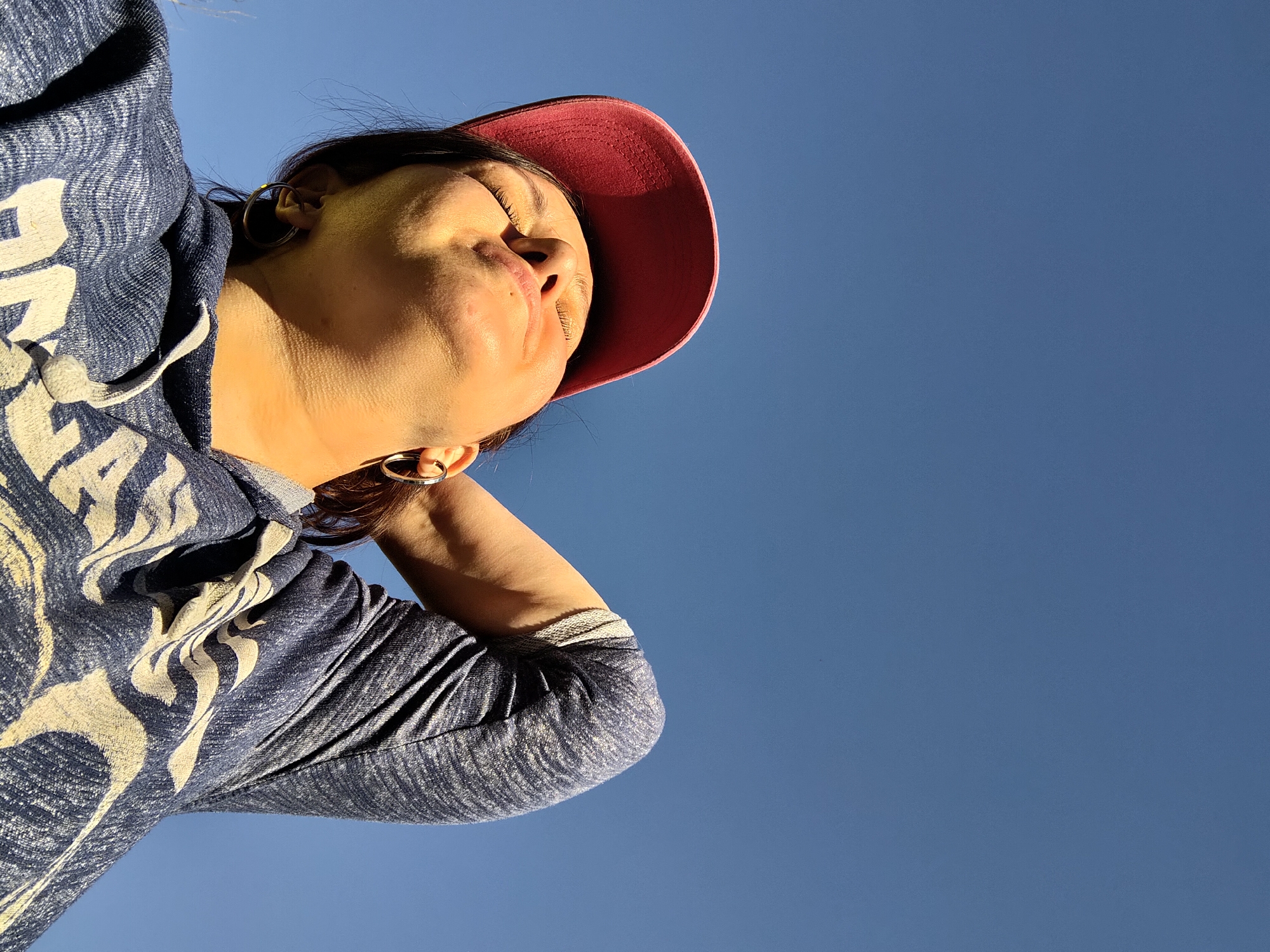 A woman is looking up toward the sky. She is wearing a maroon baseball cap and a blue hooded sweatshirt with a white pattern. Her head is tilted back, with her eyes closed. She has her hand behind her head. The background is a clear blue sky. There is no evidence that the photo was taken in Markovo, Russia.