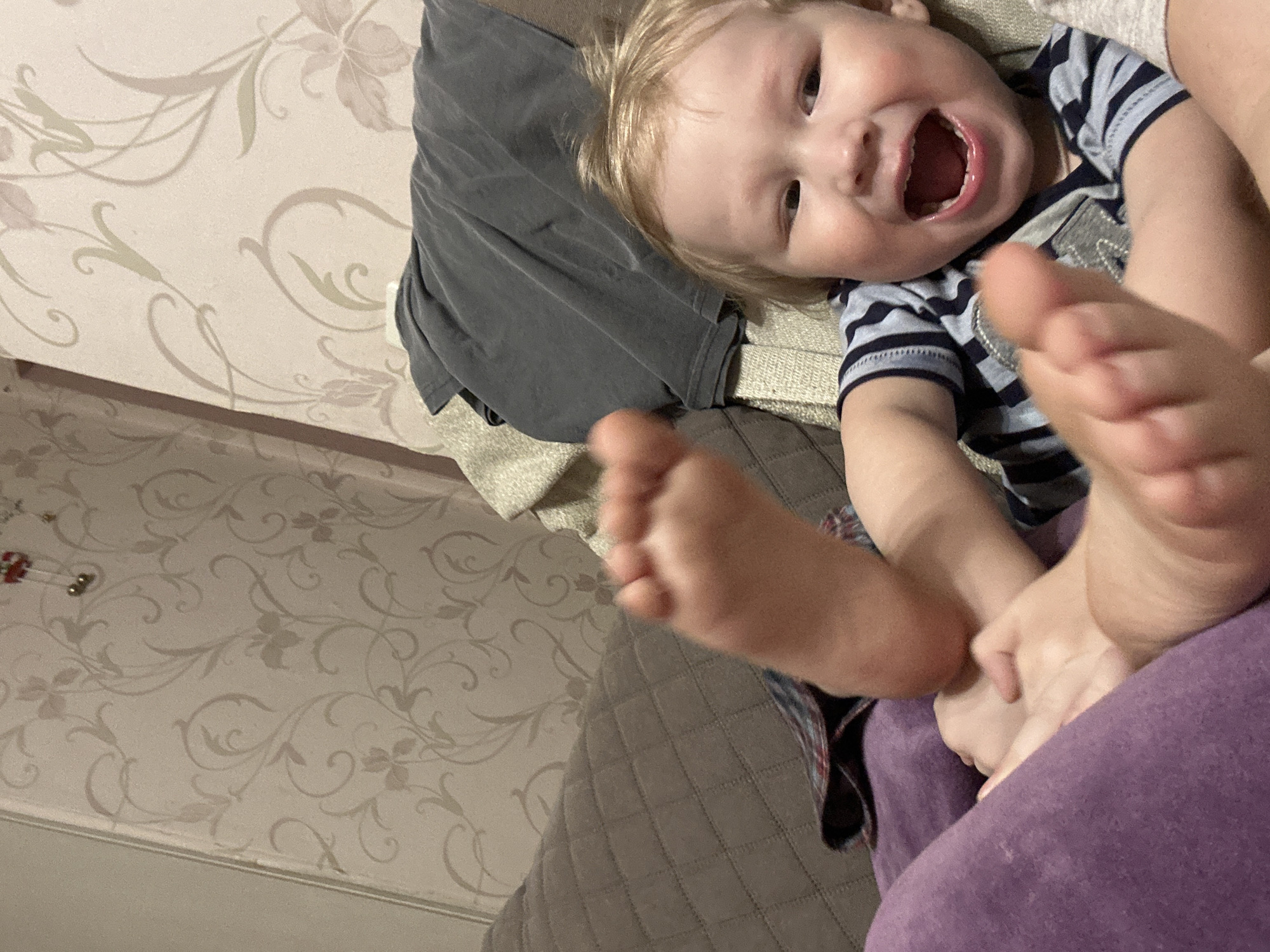 A baby is lying on a surface, possibly a bed or couch. The baby has blonde hair and is wearing a blue and gray striped shirt. The baby is smiling with their mouth open, and their feet are raised in the air. A gray t-shirt is also visible on the surface. The background includes a wall with a floral pattern. The location of Votkinsk, Russia cannot be inferred from this image.