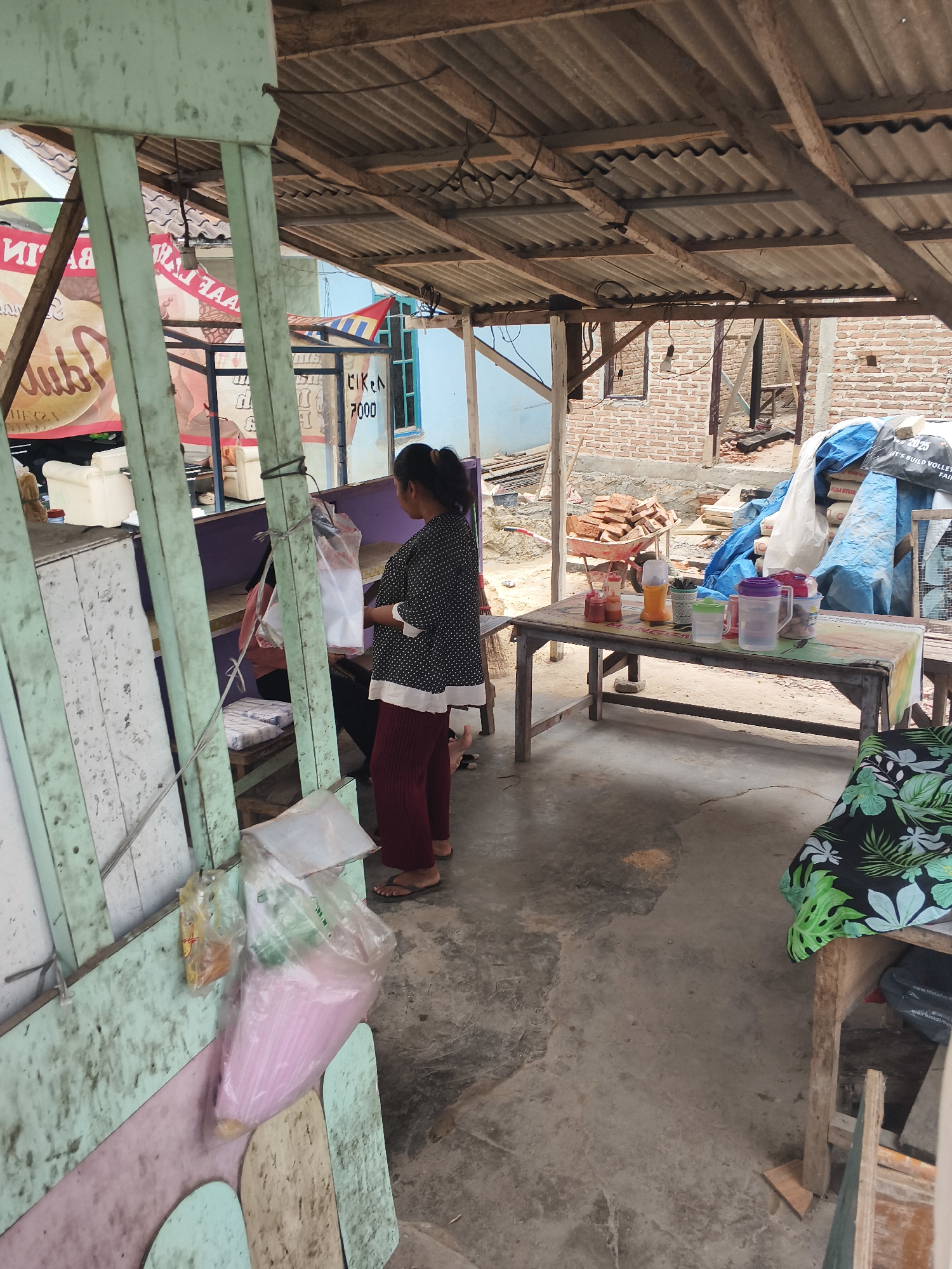 An exterior shot, likely in Bandar Lampung, Indonesia, captures a rustic, open-air market stall. The stall's roof is constructed of corrugated metal supported by wooden beams, suggesting it's a temporary or semi-permanent structure. The ground is concrete, and the scene is cluttered with various items, hinting at ongoing construction or renovations in the background, with exposed brick walls visible.
A woman, wearing a dark polka-dot top and maroon pants, stands near the stall, holding a plastic bag. Her stance suggests she might be a vendor or a customer. Another person is partially visible behind her, also appearing to be interacting with the stall.
In the foreground, a wooden table is laden with various objects, including plastic bottles of condiments (likely ketchup and chili sauce), a blender, cups, and a large pitcher of water, indicating it's set up for selling food or drinks. In the background, a wheelbarrow full of bricks and large blue tarpaulins suggest construction activity. The stall itself is made of weathered, painted wood, with some signs visible in the background that appear to be advertising or pricing, including one with the text "CIK EN 7000". The overall impression is one of a busy, functional, and somewhat unkempt marketplace environment.