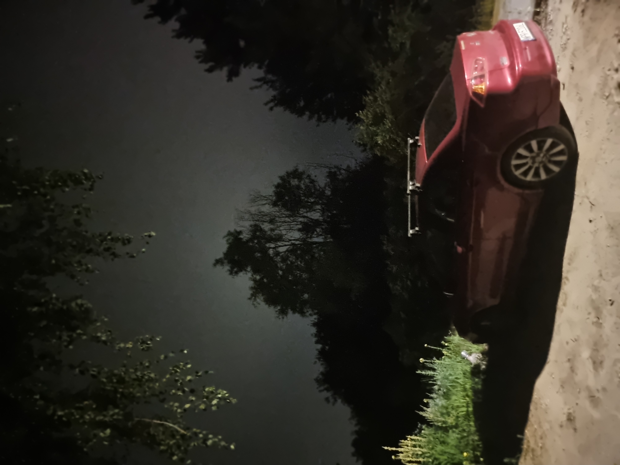 An exterior, low-angle, medium shot captures a red sedan parked on a dirt path at night in Szczaki, Poland. The vehicle, facing left, has a roof rack and a license plate reading "WPP 5Z04". Overgrown green vegetation and trees form a dark backdrop against a hazy, illuminated sky. The foreground shows a rough, sandy surface with scattered debris. The lighting suggests an outdoor, possibly rural, setting with ambient light from an unseen source.