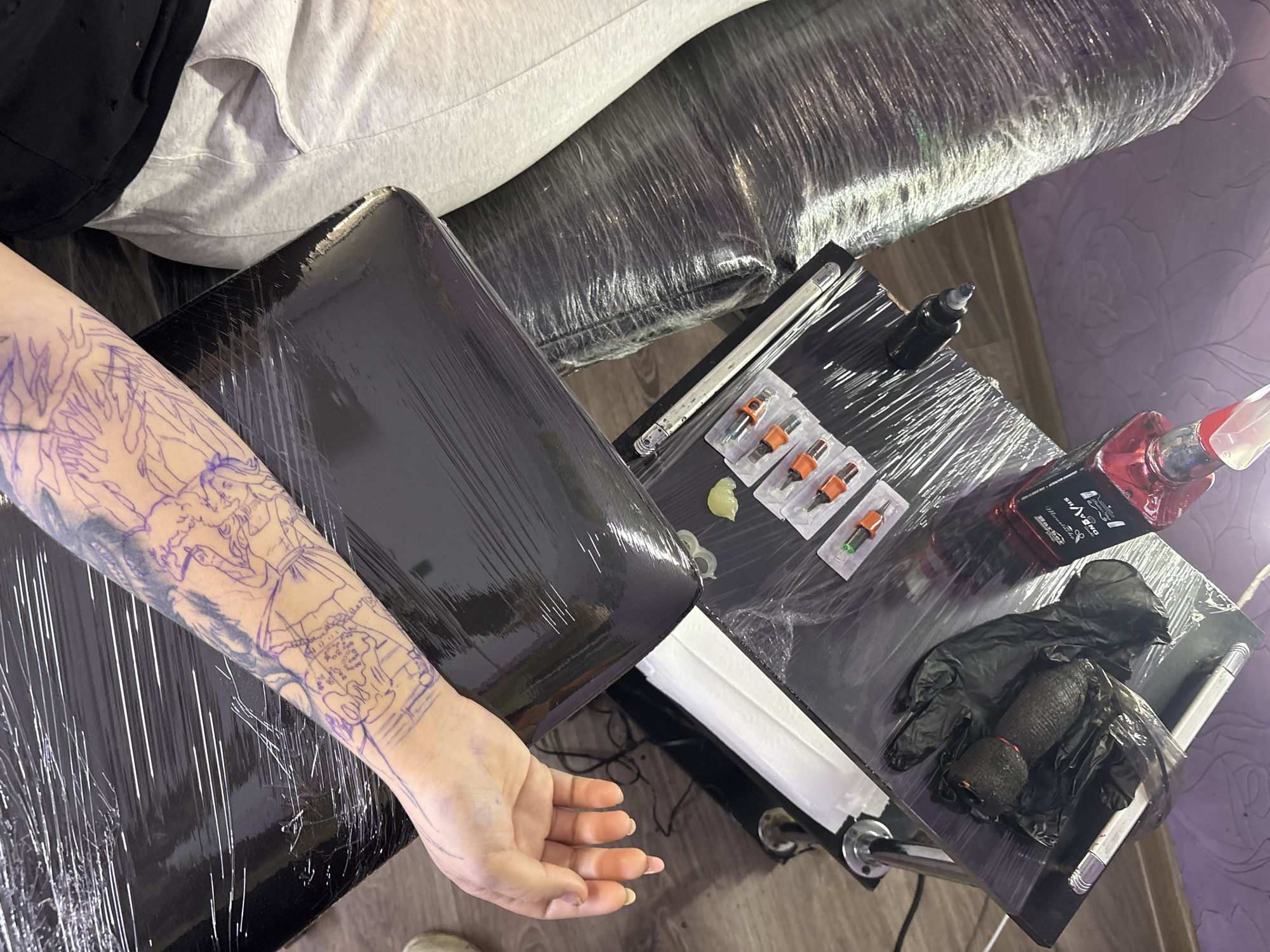 The image depicts a close-up view of a person's left forearm being prepared for a tattoo. The arm is covered with a purple stencil design, and the surrounding area is draped with black plastic wrap. On a black surface to the right of the arm, several tattoo cartridges in plastic packaging, a small amount of green ointment, a bottle of red tattoo ink, black disposable gloves, and a tattoo machine wrapped in plastic are visible. The person's hand, with neatly manicured nails, rests on the black draped surface. The background shows a wooden floor and part of a person's gray sweatpants and black t-shirt. The scene suggests a tattoo artist's workspace, likely a studio. Without further information or visual cues, it is impossible to determine the specific location in Almaty, Kazakhstan.