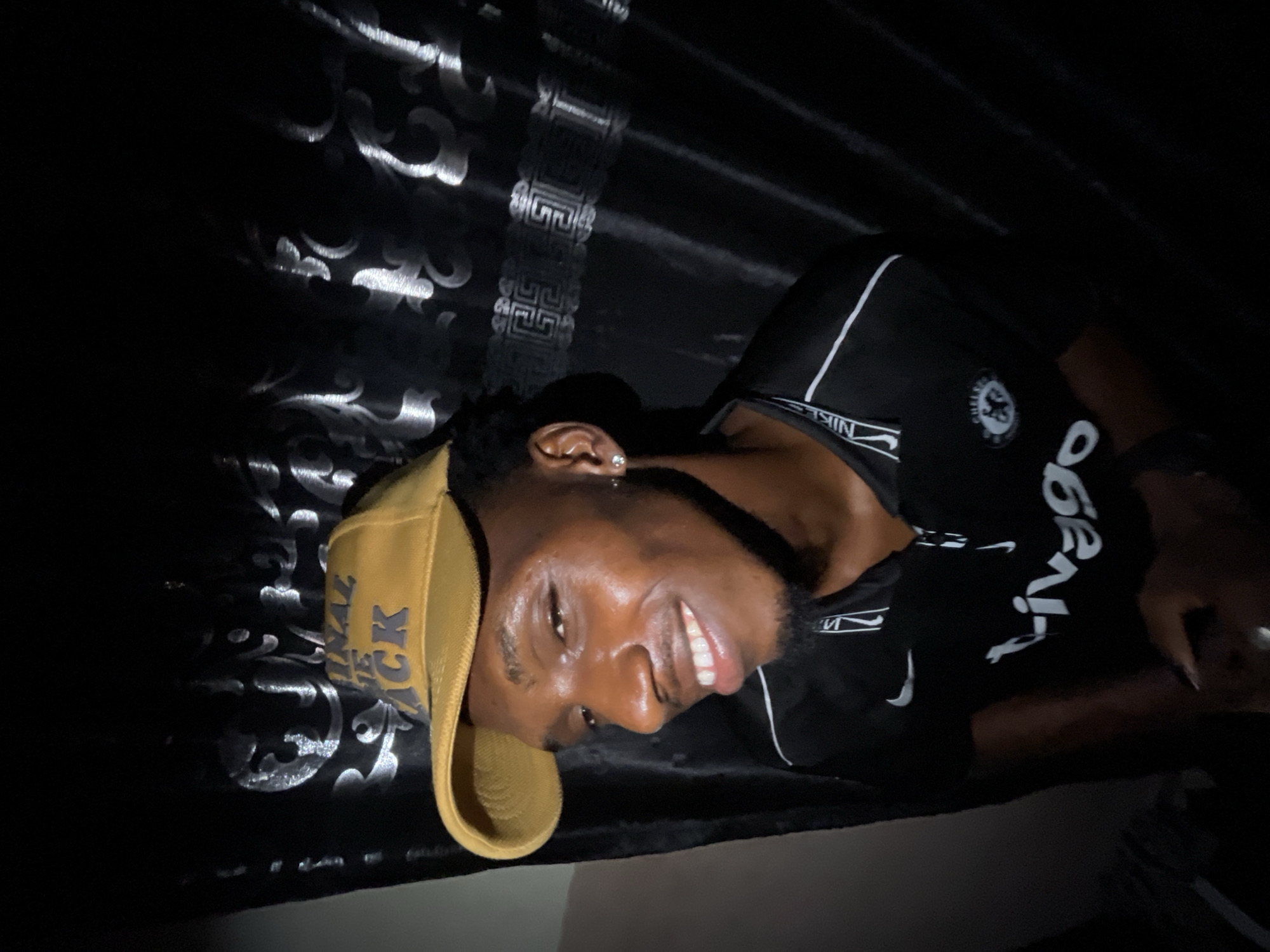 A dark-skinned man wearing a tan baseball cap, a black Chelsea soccer jersey with white lettering, and a small earring is lying on a black surface with a silver paisley pattern. The location and city of Lokoja, Nigeria are not determinable from the image.