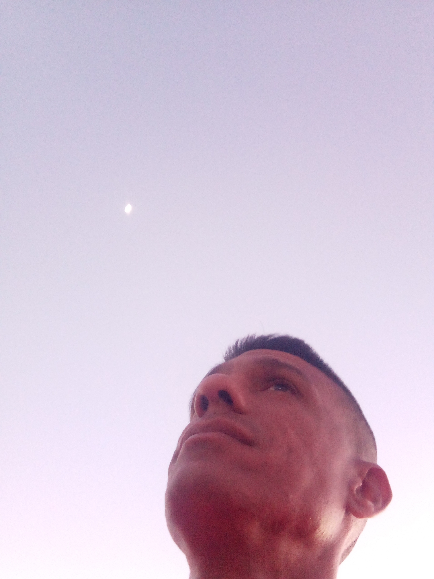 A low-angle shot shows a person looking up at a light or the moon in a light purple sky. The person has short dark hair and is Caucasian. The location is Unknown, Unknown.