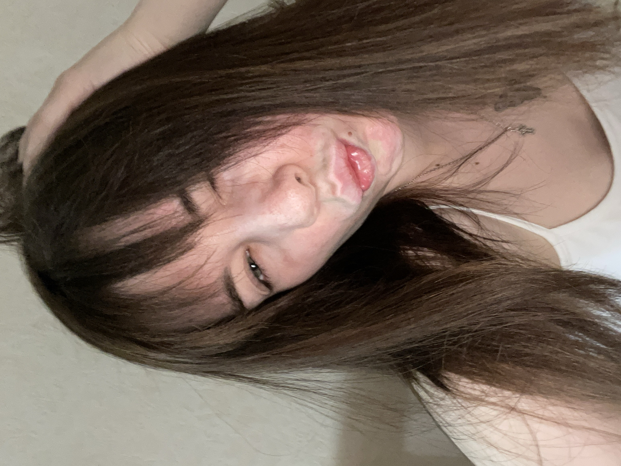 A young woman with long, dark brown hair is positioned diagonally in the frame. Her head is tilted towards the left, and she is partially obscured by her hair, which falls across her face. Her left eye is partially visible, and she has a slight smirk. Her hand is touching her hair above her head. She wears a white tank top. A silver chain necklace is visible on her neck. There appears to be a tattoo on the upper right side of her chest. The background is a plain, light-colored wall.
There is no discernible information indicating the location is the city of Chaikovsky, Russia.