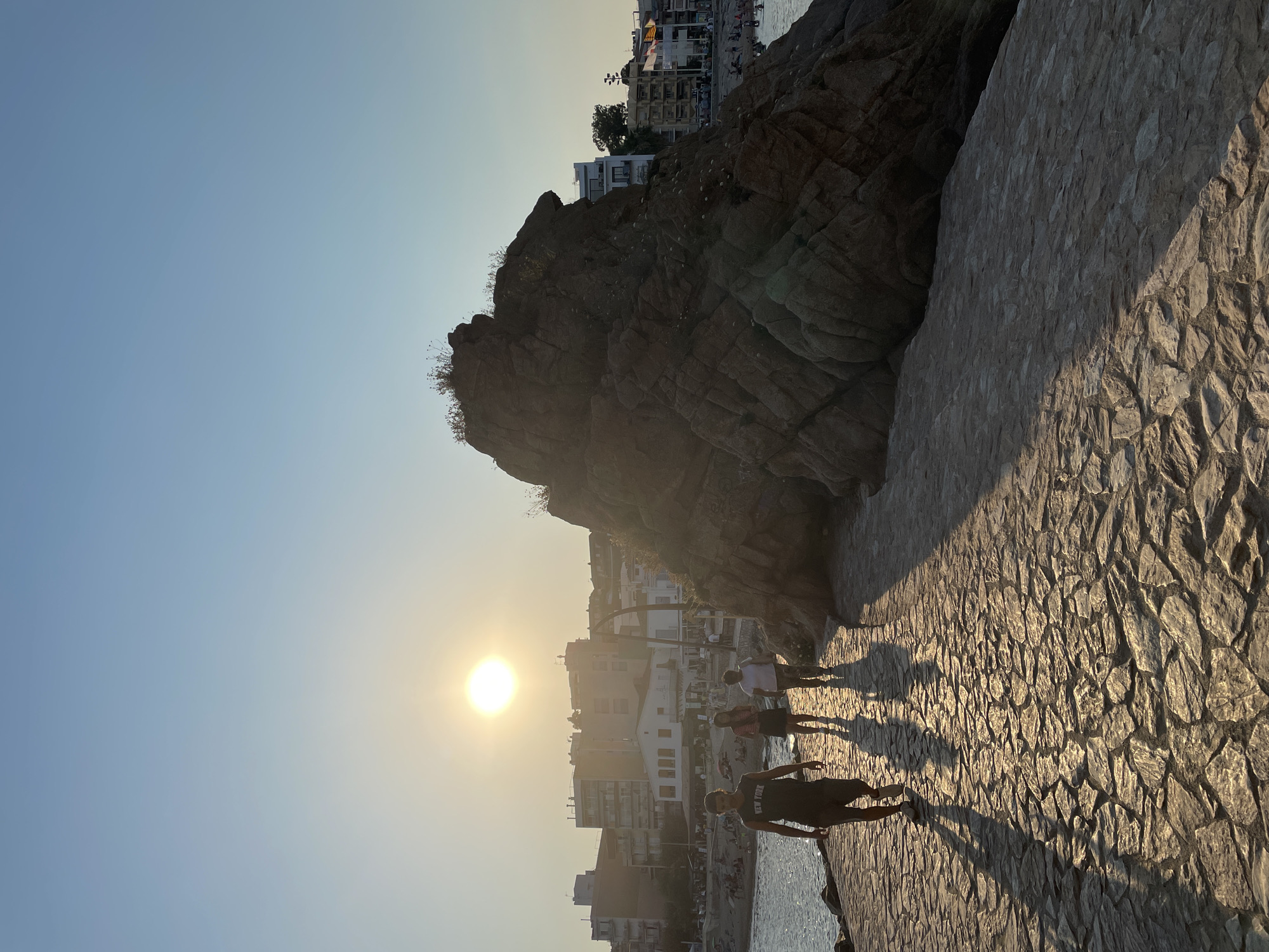 The image depicts a paved walkway along the coast in Blanes, Spain, during what appears to be late afternoon or early evening. The sun is low in the sky, casting long shadows across the stone pathway. Several people are walking on the path, silhouetted against the bright sunlight. In the background, buildings line the coast, and a large rock formation dominates the right side of the frame. The sea is visible to the left of the walkway, and several people can be seen in the water. The overall scene conveys a sense of a leisurely stroll on a warm day in a coastal town.