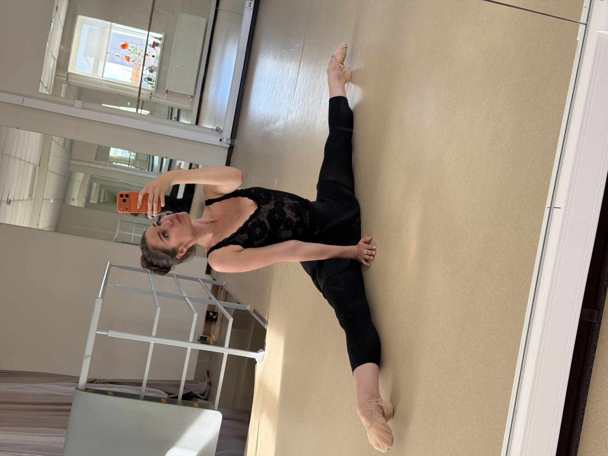 A woman is performing a split in a dance studio, holding her phone to take a mirror selfie. She is wearing a black leotard with lace detailing and black leggings, with ballet slippers on her feet. The studio has a large mirror on one wall, reflecting the woman and the room. Sunlight streams in from a window, illuminating the space. In the background, a white radiator and a shelf with decorative items are visible. The floor is a neutral beige. The scene suggests a dedicated practice session, with the woman focused on her form.