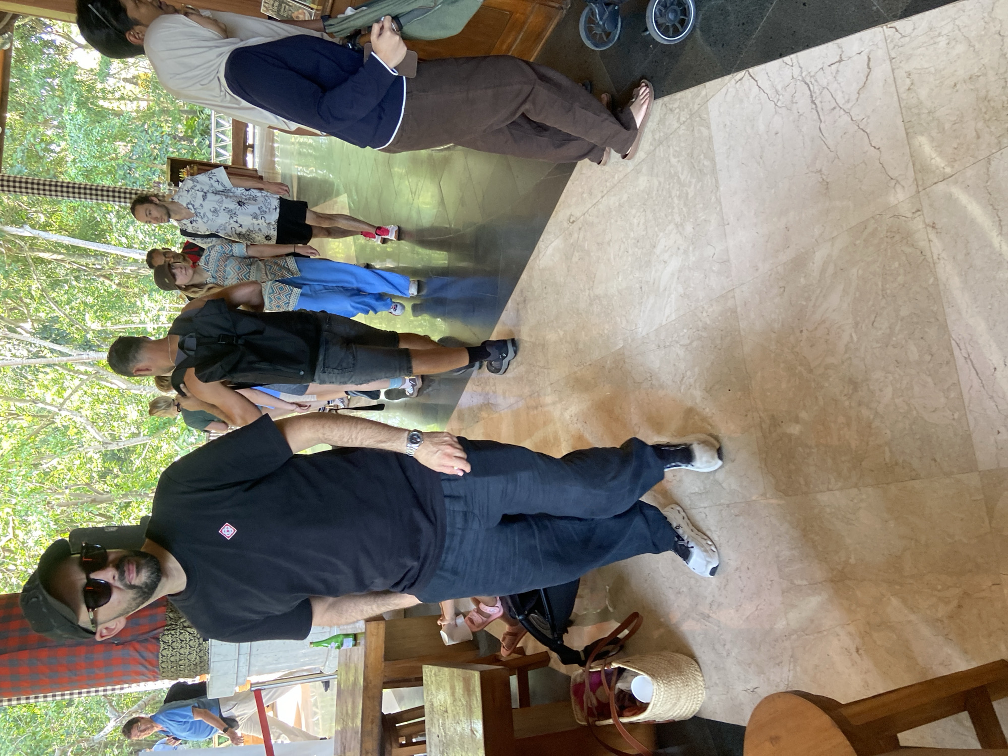 In Ubud District, Indonesia, a group of people are walking through a covered walkway with a tiled floor. The man in the foreground is wearing a black t-shirt, black pants, a baseball cap, and sunglasses. He has a beard and is looking to his left. To his right, a woman in a blue headscarf and dark blue shirt is walking, carrying a baby stroller. Further back, a man with a backpack is walking away from the camera, and other individuals are visible in the background, some wearing casual clothing. The environment appears to be an outdoor or semi-outdoor space with lush green foliage visible through the openings. Red and black checkered fabric is hanging on a pillar behind the man in the foreground. There are wooden tables and chairs to the left of the frame, and a woven basket is on the floor.