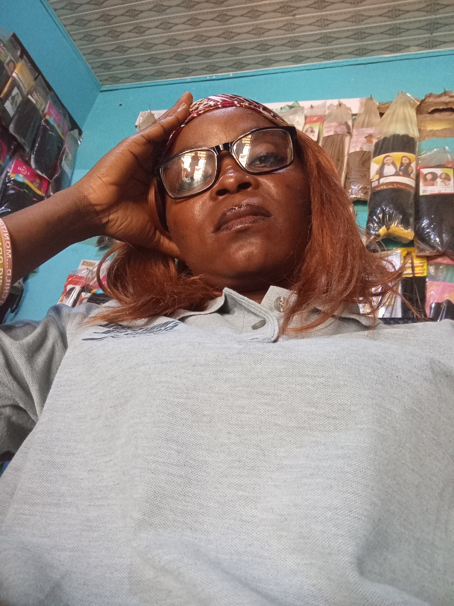 The image shows a woman in a store. The woman is wearing a grey polo shirt, glasses, and a headscarf. She has red hair and is resting her hand on her forehead. Behind her, shelves are stocked with hair products, suggesting the store is a beauty supply store. The store appears to be located in the city of Jalingo, Nigeria.