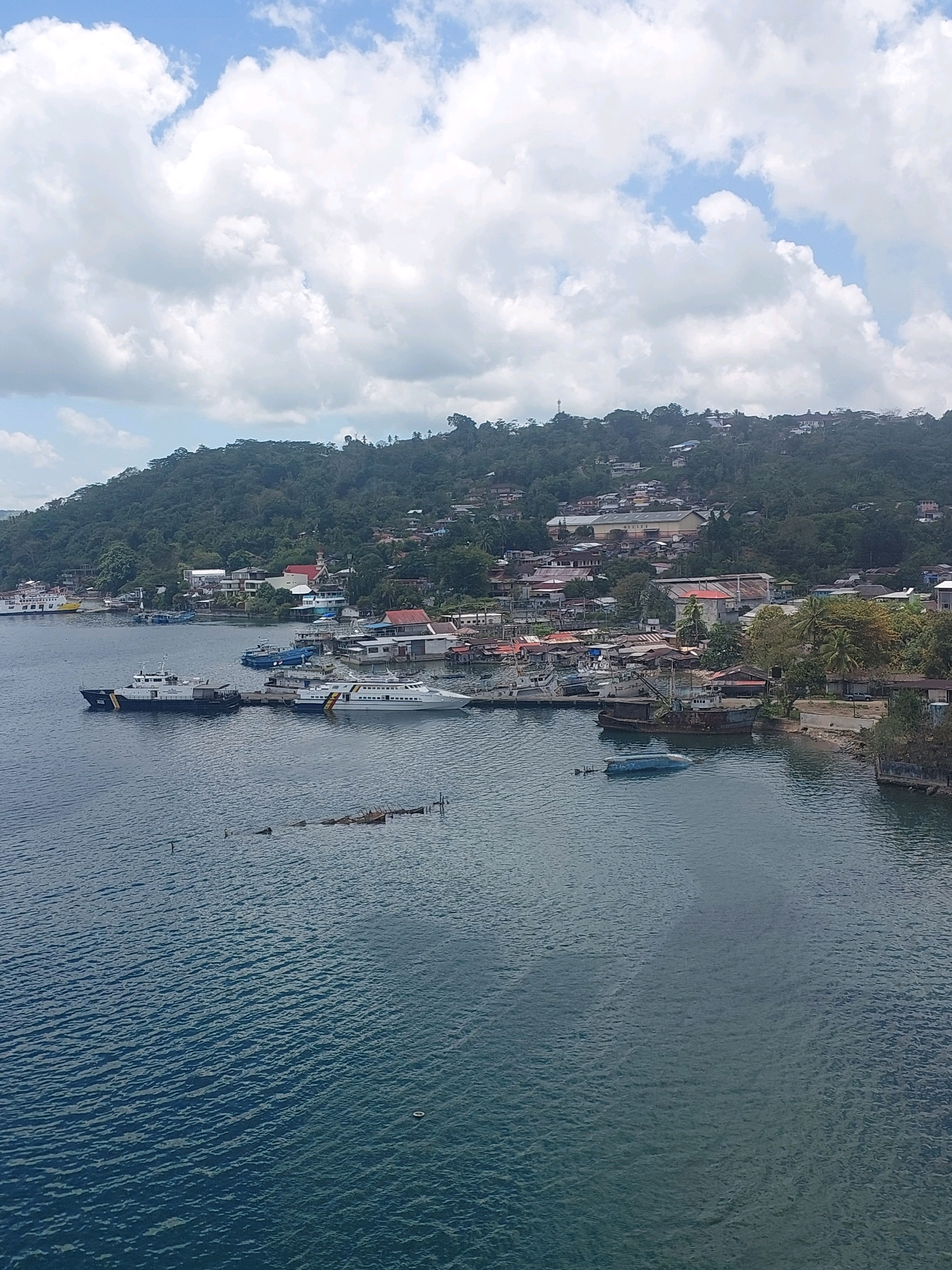 An aerial view captures the waterfront of Ambon City, Indonesia, with the turquoise waters of the bay dominating the foreground and middle ground. Numerous boats of various sizes are moored or docked along the shore, including several white passenger ferries, a dark-hulled patrol boat, and older, rusted vessels. Buildings of different architectural styles, some with red roofs, are clustered on the lush green hillside that rises steeply behind the waterfront. The sky is a mix of bright blue and dramatic white clouds. The overall scene depicts a bustling coastal community with ongoing maritime activity.