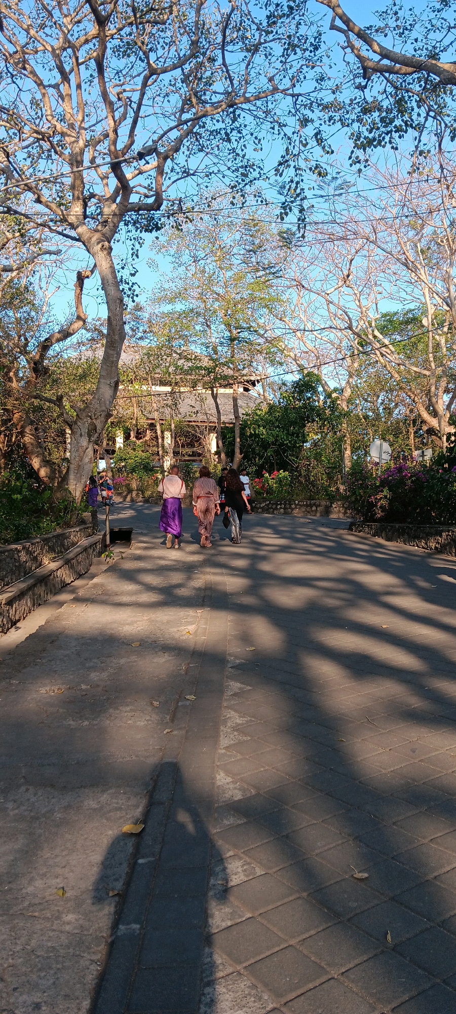In Kuta Selatan, Indonesia, a paved walkway meanders through a park-like setting with mature trees casting dappled shadows. Three individuals are walking away from the viewer down the path. The first person, on the left, wears a long, flowing purple garment. Next to them, a second person is clad in a striped, patterned outfit. A third person, on the right, wears dark clothing. Further ahead, another individual in dark clothing is visible. The background features lush greenery, flowering plants, and a building with a distinctive roofline, likely a temple or cultural structure, suggesting a destination within the area. The sun is low in the sky, creating long shadows across the walkway and illuminating the scene with a warm, golden light.