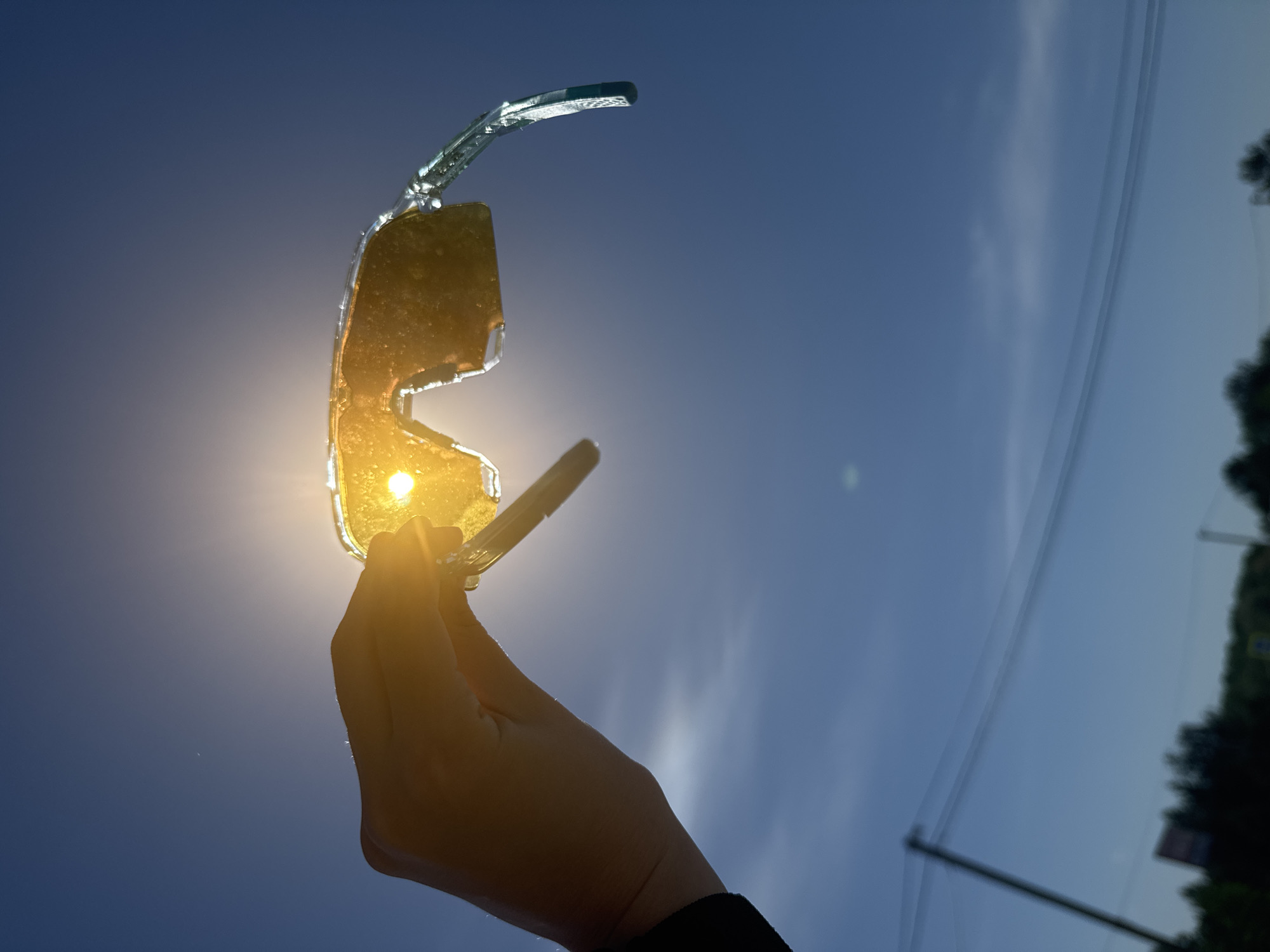 The image depicts a hand holding sunglasses up to the sun. The sky is blue and clear, and there are trees and power lines visible in the background. The location is likely outdoors. The city is unknown.