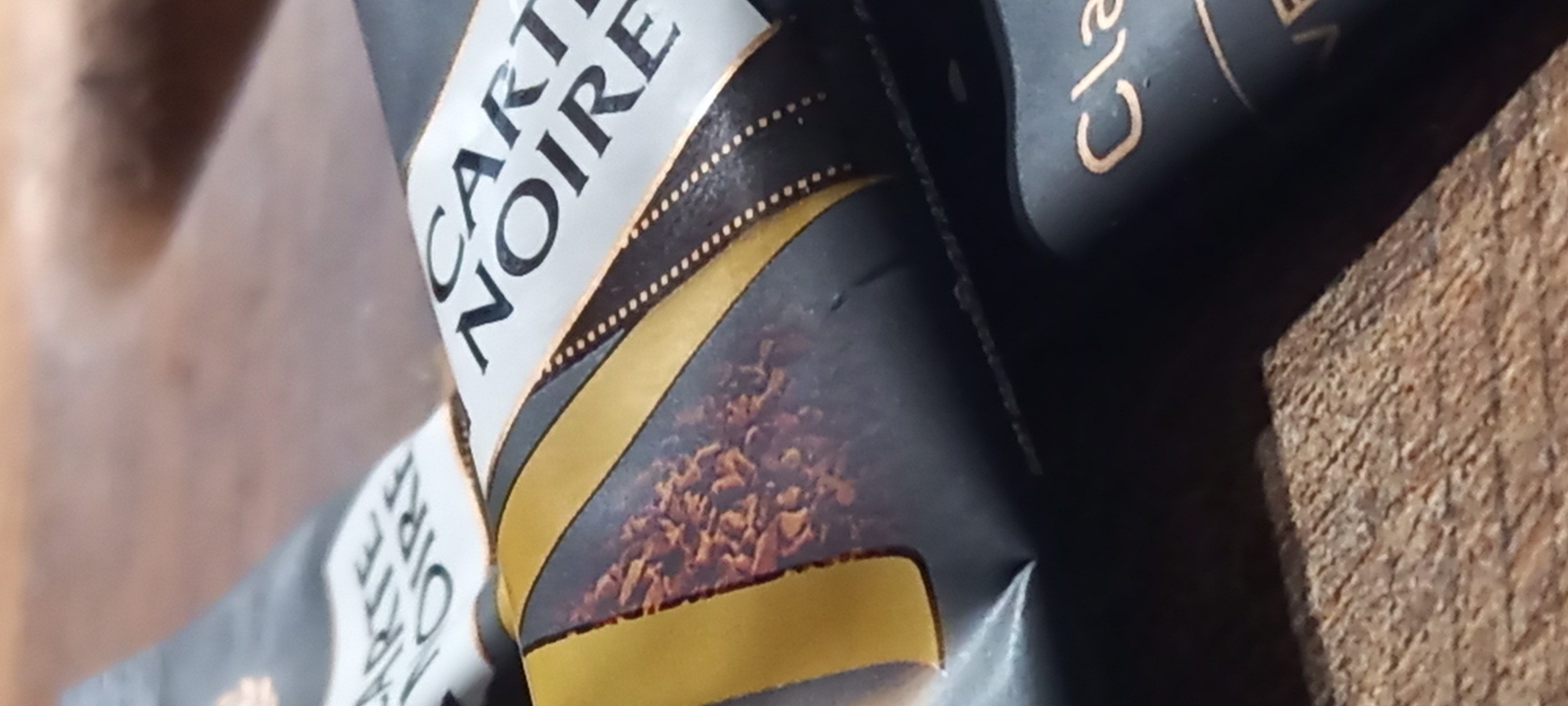 A close-up, diagonally oriented shot features a black coffee packet, likely a sachet or pouch, resting on a wooden surface. The main subject is the coffee packaging, which has a prominent white and gold label. The brand name "CARTE NOIRE" is clearly visible on the white section of the label, with black serif typography. Below this, a golden band separates the text from an image of dark brown, granular coffee grounds. Another part of the packaging, to the right, partially shows the word "Cla", possibly indicating a type like "Classique". The background consists of a light brown, grainy wooden surface, visible in the upper left and lower right corners of the frame. The lighting is soft, suggesting an indoor environment. There are no people or discernible activities taking place.