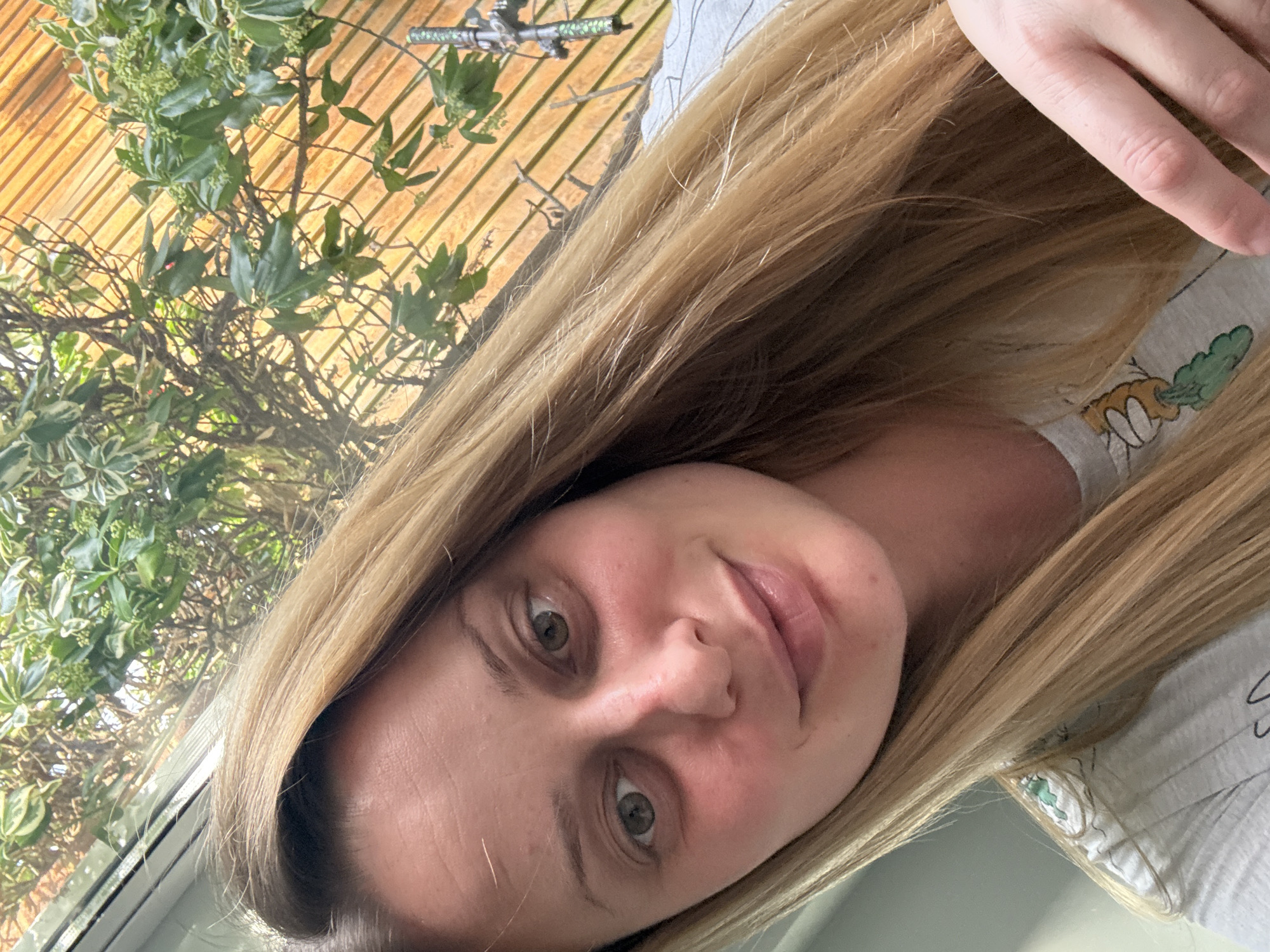 A selfie photograph shows a blonde-haired woman with fair skin, green eyes, and a slight smile. She is wearing a light-colored t-shirt with cartoon-like printed motifs. Behind her is a window revealing a verdant outdoor scene with a wooden fence-like structure partially obscured by foliage. The location is likely indoors, judging by the window frame, but there are not enough details to definitively pinpoint a specific area of Hazlemere, United Kingdom. The time of day appears to be daytime due to the brightness of the background.