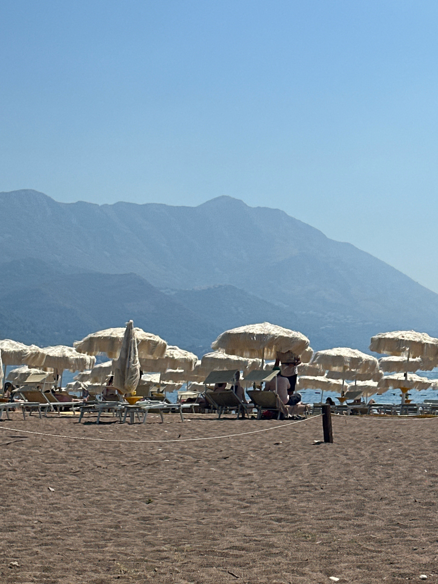 This image depicts a sun-drenched beach with numerous beige umbrellas and empty lounge chairs set up on dark sand. In the background, the calm blue water of the sea is visible, meeting a hazy mountain range under a clear, pale blue sky. Several people are scattered across the beach, some reclining on lounge chairs under umbrellas, while others are standing or walking. The scene appears to be from the city of Boreti, Montenegro, given the characteristic mountain landscape that borders the coast. The overall atmosphere is one of a leisurely summer day at the seaside.