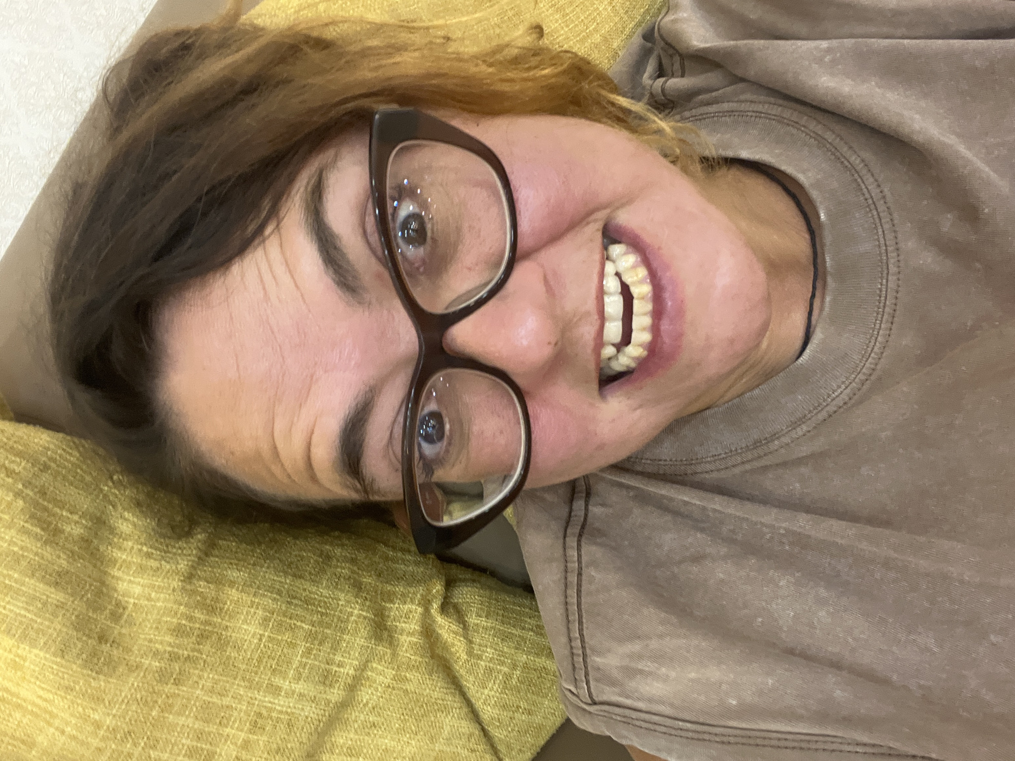A woman with brown hair, wearing large-rimmed glasses and a brown t-shirt, smiles widely, exposing her teeth. She is lying down, her head resting on a yellow, textured surface, likely a cushion or sofa. The background is a neutral-colored wall. The image was taken in Nha Trang Ward, Vietnam.