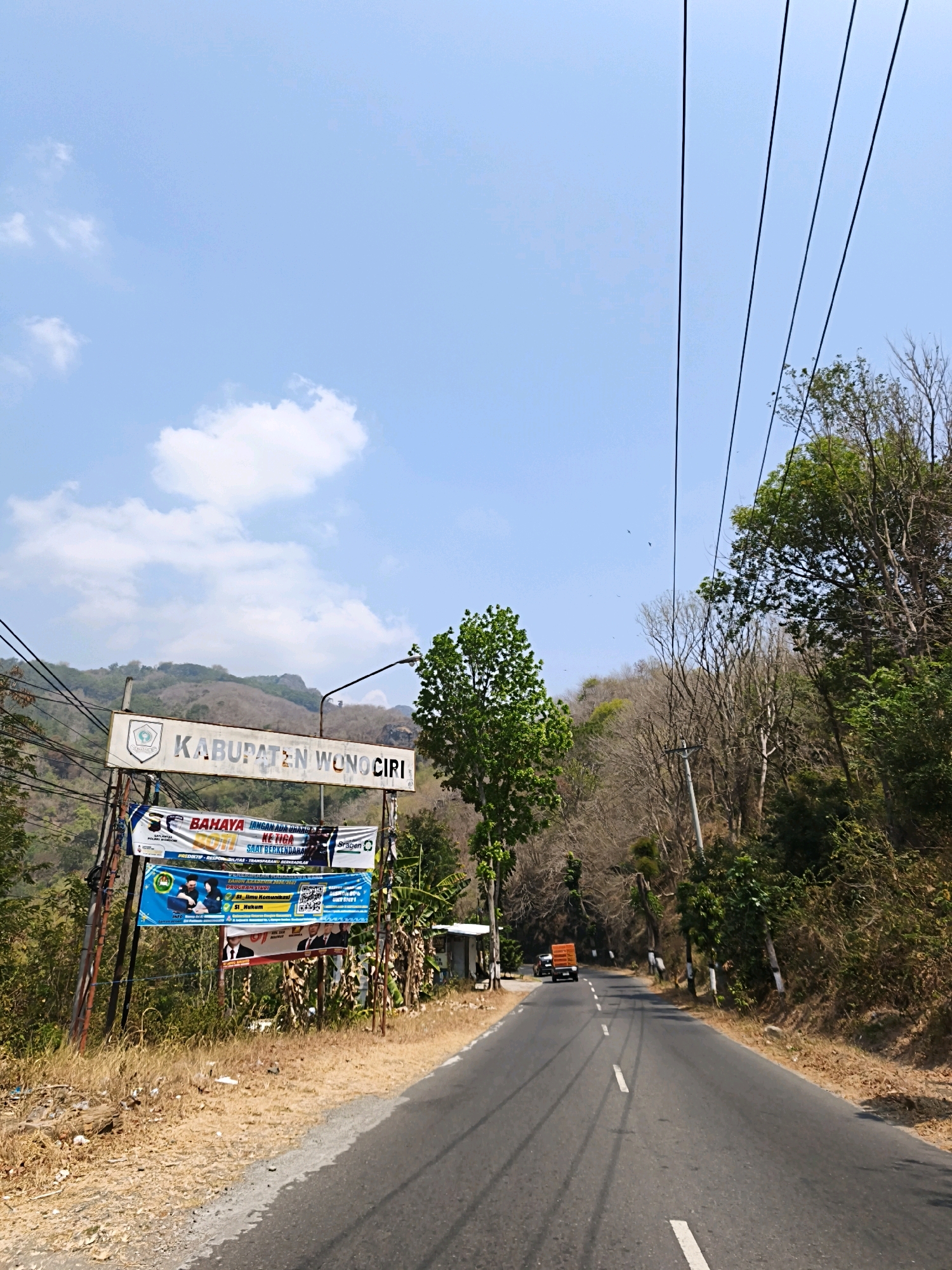The image depicts a road in Wuryorejo, Indonesia, on a clear, sunny day with a few clouds in the sky. A signpost on the left side of the road displays "KABUPATEN WONOCIRI," indicating the administrative region. Below this, several banners are hung. One banner features text in Indonesian, including "BAHAYA" (danger) and "JANGAN ADA DURHAKA KE TIGA SAAT BERKENDARA" (no disrespect to the third time while driving). Another banner shows an image of people in suits and text related to "PROGRAM STUDI" and "SI_Ilmu Komunikasi" along with a QR code. The road itself is asphalt and appears to be in good condition, with white lines marking the lanes. On the right side of the road, lush green trees and dry vegetation are visible, characteristic of a rural or hilly environment. In the distance, a truck with an orange container is traveling away from the viewer. Power lines stretch across the frame, adding to the natural and somewhat utilitarian landscape. The overall scene suggests a typical day in the Indonesian countryside, with a focus on signage and the road infrastructure.
