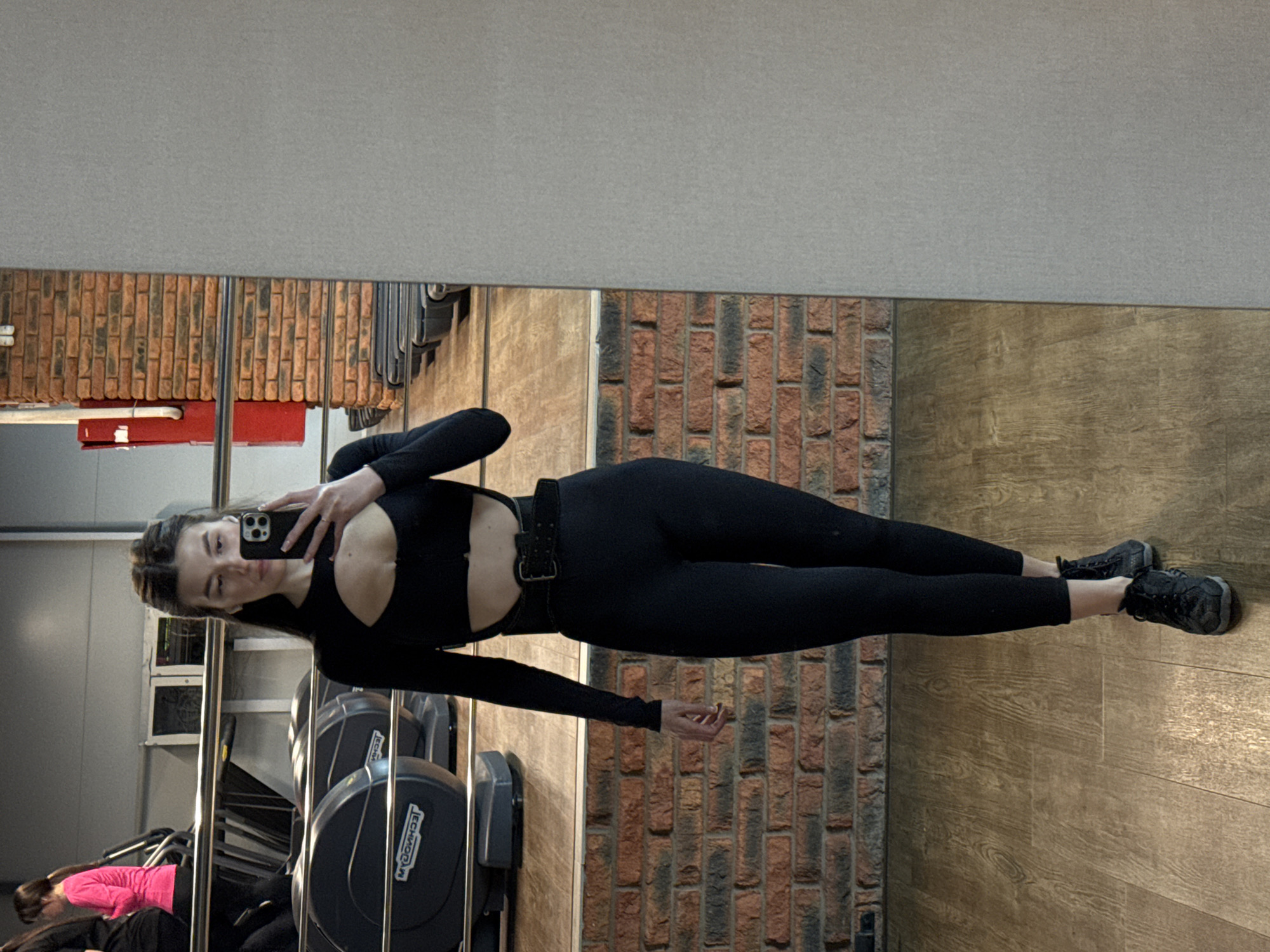 The image features a young woman, likely in her 20s, taking a mirror selfie in what appears to be a gym or fitness center. She stands upright, facing her smartphone which she holds up to capture her reflection.
The woman is wearing a fitted, long-sleeved black athletic top with a distinctive cutout on the chest, black leggings, and black athletic shoes. A black belt with a silver buckle is cinched around her waist. Her brown hair is styled away from her face, and she maintains a neutral expression while looking at her phone.
The background visible in the mirror reflection includes a textured brick wall with a mix of red, brown, and grey tones, alongside a segment of wooden paneling or flooring. Above the brickwork, the wall is a plain light grey. The floor beneath her feet is also made of light brown wooden planks.
Various pieces of gym equipment are reflected, particularly in the lower left, including machines that resemble treadmills or elliptical trainers. The brand name "TECHNOGYM" is clearly visible in white lettering on the black surfaces of some of this equipment. Another person, wearing a bright pink top and black bottoms, is partially seen in the reflection, engaged with one of the gym machines. The scene depicts a typical indoor fitness environment, consistent with a gym in Chaikovsky, Russia.