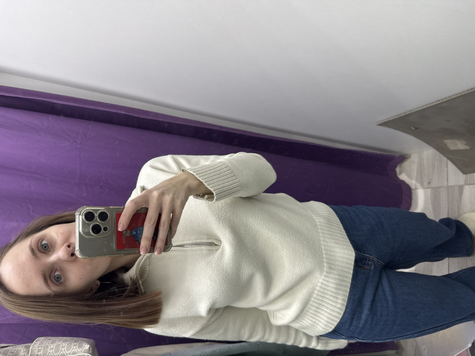 An adult female, appearing to be in her late twenties to early thirties, with light brown, shoulder-length hair and blue eyes, is depicted taking a mirror selfie. She wears a cream-colored, textured knit sweater with a half-zip collar and ribbed cuffs and hem, paired with dark blue jeans and light-colored slippers or shoes. Her left hand holds a smartphone, equipped with a clear case and a red card holder displaying a graphic. A thin, gold-colored bracelet is visible on her right wrist. The setting is consistent with a retail changing room, featuring a purple fabric curtain on the left side of the reflection and a plain white wall on the right. A light-colored bag with a repeating pattern and another garment are partially visible behind the curtain. The floor is light-colored and patterned, and a grey panel or object lies against the white wall at the bottom right. No specific identifying markers are present within the image to pinpoint a precise location within Perm, Russia.