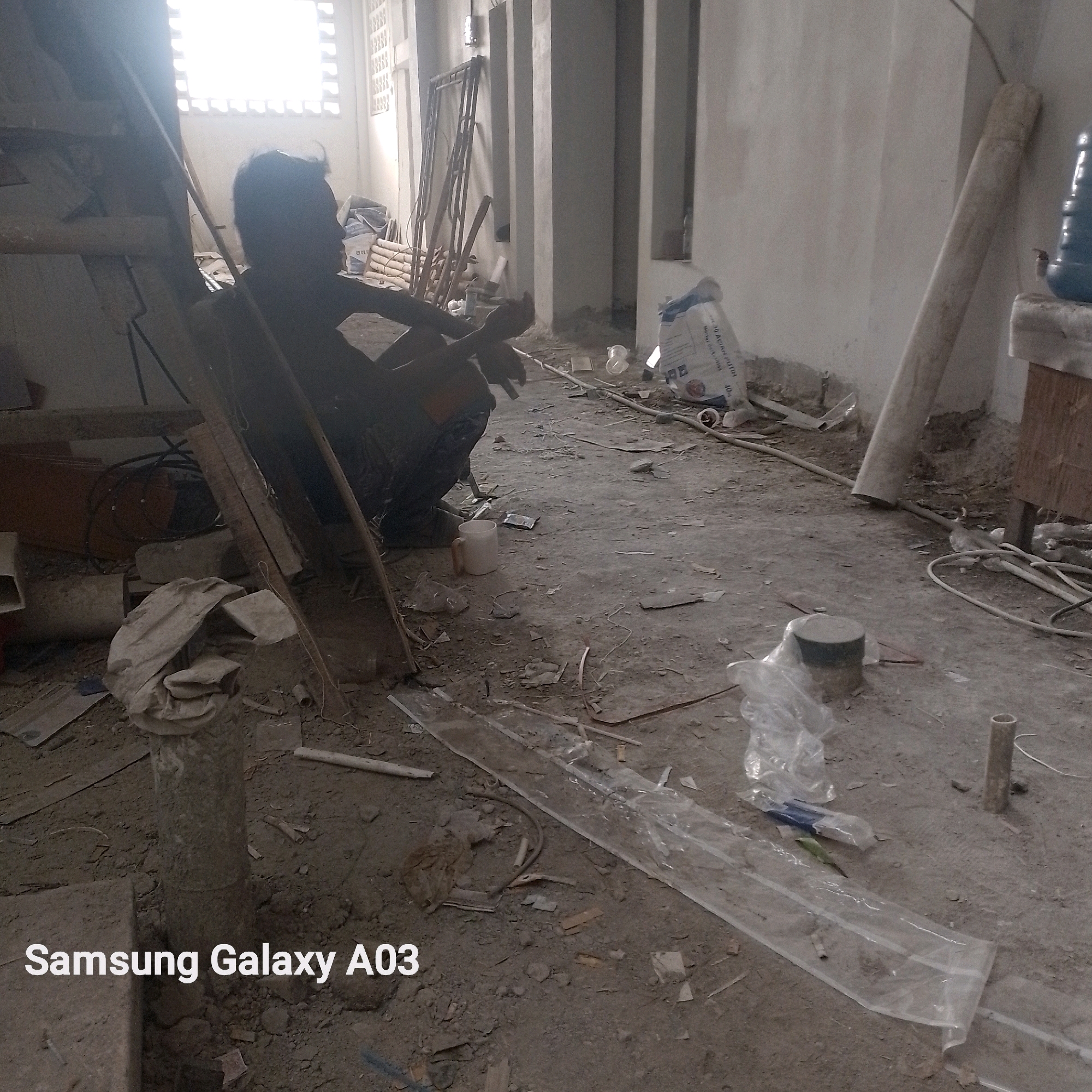 A dimly lit construction site in Indramayu, Indonesia, is the setting for this image. A lone individual, silhouetted against a bright window, is seated on the ground, appearing to be engaged in some activity with their hands. The floor is covered with dirt, debris, and construction materials, including pipes, bags of what appears to be cement, and plastic sheeting. The walls are bare and unfinished, and the overall impression is one of ongoing construction or renovation. The Samsung Galaxy A03 watermark is visible in the lower left corner of the image.
