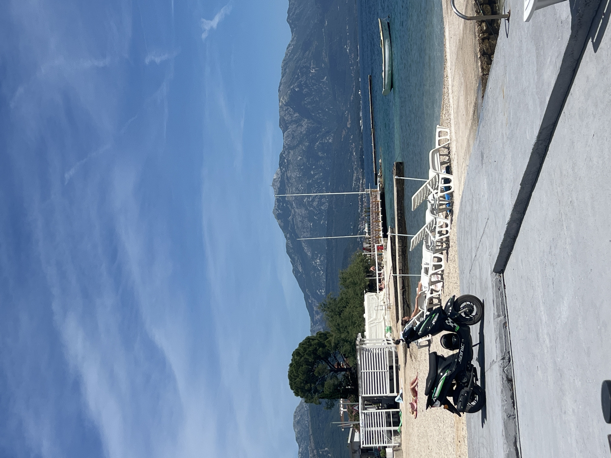 The image captures a view of a beach area, possibly in Baošići, Montenegro, given the presence of mountains in the background and the coastal setting. A body of water, likely the sea, stretches into the distance, with a single boat visible. A pier extends into the water. The beach area is populated with white lounge chairs. A moped rests on a paved walkway alongside the beach. People are seen relaxing on the beach. The sky is clear with some clouds. A tree is present near the beach area. The overall impression is that of a summer scene at a resort or beach location.