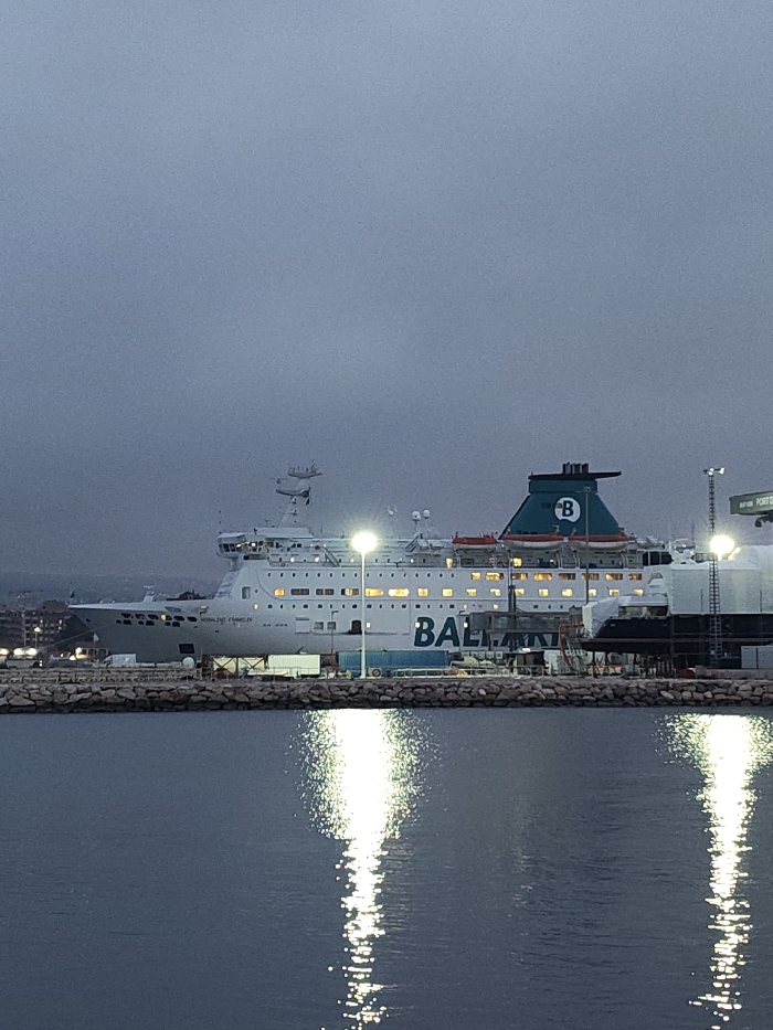 The image depicts the white passenger and vehicle ferry "ROSALIND FRANKLIN," operated by Balearia, moored at a pier within the port of Dénia, Spain. The scene is illuminated by artificial light sources under low ambient light conditions, suggesting either dusk or dawn. The ferry's interior windows glow, and multiple bright pier-mounted lights cast prominent, elongated reflections onto the calm, dark water in the foreground. The vessel's name is visible on the bow, and the Balearia logo is displayed on the hull and on the vessel's green funnel. The pier itself features a rocky breakwater, behind which industrial port equipment, including what appears to be a portion of another vessel undergoing maintenance or a dry dock structure, is present. Distant city buildings and topographical features are faintly discernible in the background. No individuals are clearly visible within the frame.