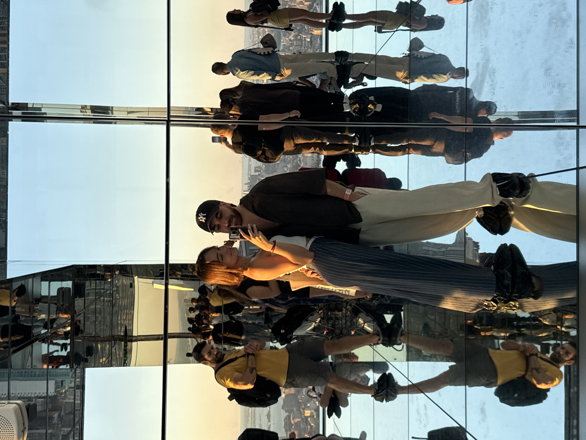 The image captures an indoor observation deck featuring a striking, highly reflective environment, where mirrored surfaces create an illusion of infinite space. The photograph is oriented vertically, but depicts people standing upright on what appears to be a mirrored floor and against mirrored walls and ceiling.
In the foreground, a man with a beard wearing a black baseball cap with a white "NY" logo and a dark top is seen looking downwards. Beside him, a woman with brown hair, dressed in a white strapless top and striped pants, holds a smartphone, seemingly capturing a moment or taking a selfie, and looks towards the man.
Numerous other individuals are visible throughout the frame, primarily as reflections. These include a man in a yellow t-shirt and grey shorts carrying a backpack, a man in a light blue shirt, and a woman in a yellow dress, all multiplied and distorted by the reflections.
The setting appears to be a modern architectural attraction, likely an observation deck high above a city, consistent with locations like those in New York, USA, as suggested by the context. The sky, visible in the reflections, shows a gradient from soft blue to warm golden tones, indicative of either early morning or late afternoon sunlight. The city skyline is faintly visible in the distance, further confirming an urban environment.
Visitors are engaged in typical tourist activities, such as exploring the unique space and capturing photos or videos with their smartphones. The central couple seems to be enjoying their time together within this immersive, reflective installation. The prominent "NY" logo on the man's cap further hints at a New York location. The overwhelming use of mirrors creates a disorienting yet visually captivating experience, making it challenging to differentiate between real subjects and their mirrored images.