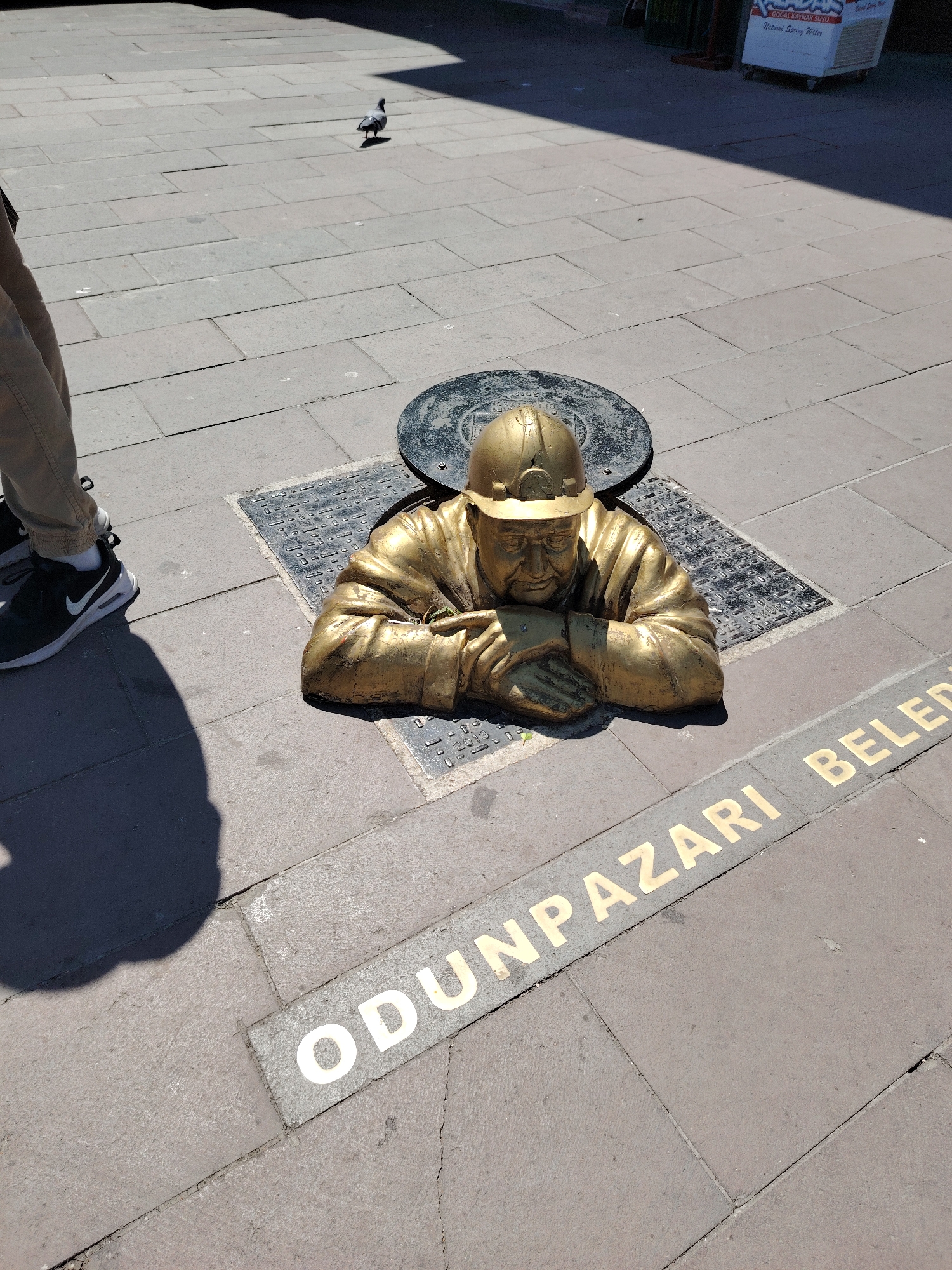 The image displays an outdoor scene on a paved urban ground, likely a public square or pedestrian area in Eskisehir, Turkey. The central focus is a striking golden-colored statue of a man, presumably a miner, appearing to emerge from a manhole cover. The statue depicts the man from the chest up, wearing a hard hat and a work jacket, with his hands clasped in front of him. The manhole cover behind the statue is dark and circular, set into a square metal frame on the pavement.
To the left of the statue, the lower legs and feet of a person are visible, wearing light-colored trousers and black sneakers with white soles and a Nike logo. A distinct shadow from this person falls across the paved ground in the foreground. Further back and to the left, a single pigeon walks on the pavement. In the upper right background, a white cooler is partially visible, featuring red lettering that reads "DOĞAL KAYNAK SUYU" and "Natural Spring Water."
The scene is bathed in bright sunlight, suggesting daytime, likely midday or early afternoon, as evidenced by the sharp, defined shadows. The ground is constructed from light gray rectangular concrete pavers. Large golden letters are embedded into the pavement in the foreground, spelling out "ODUNPAZARI BELEDİYESİ." While the full text is not entirely visible, "ODUNPAZARI BELED" can be clearly read. Odunpazarı is a district within Eskisehir. The overall impression is a unique piece of public art integrated into the city's infrastructure.