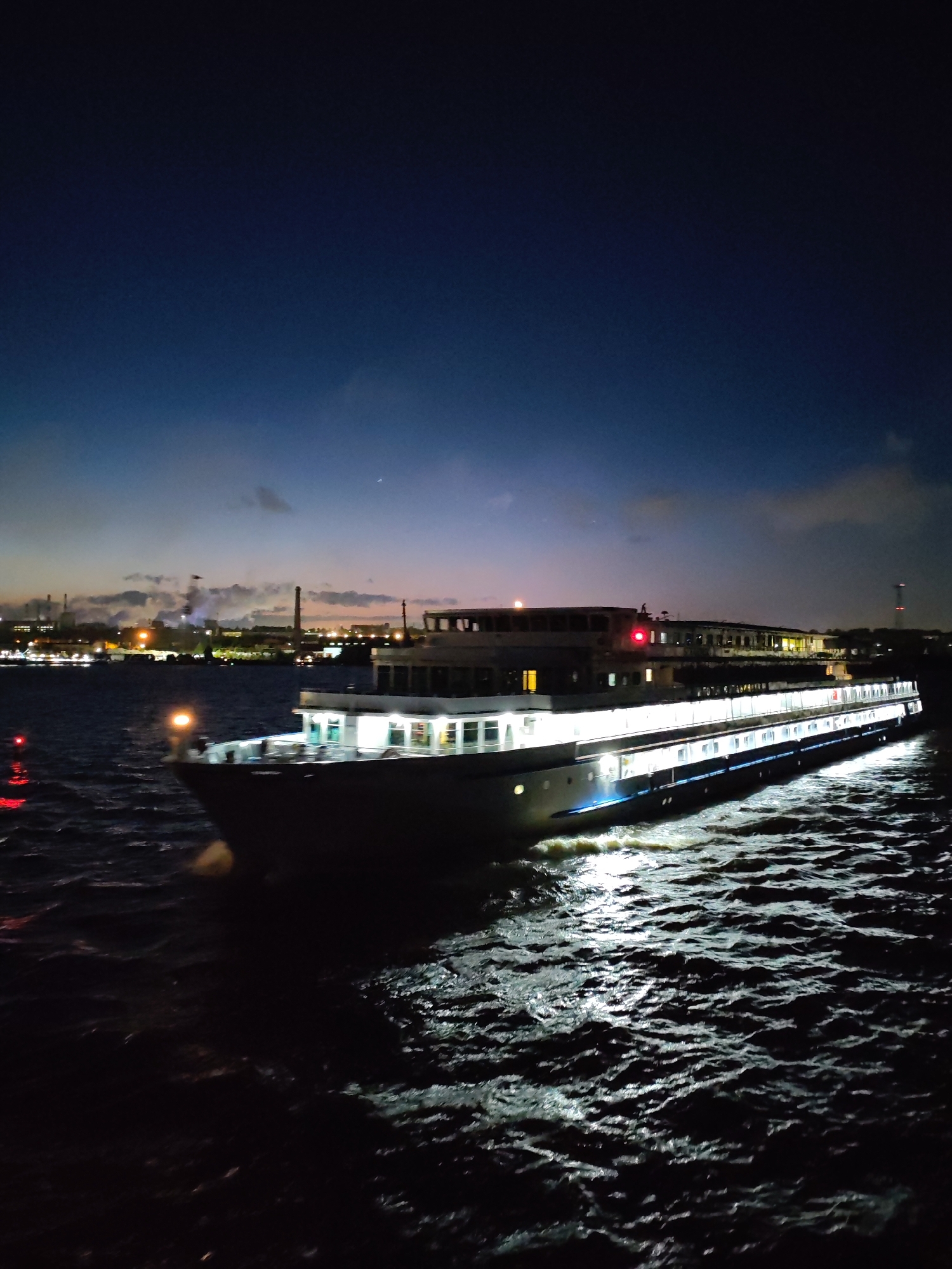 The image captures a large, illuminated cruise ship moving on a body of water, likely a river or a large lake. The lighting suggests it is nighttime, with the sky transitioning from a dark blue to a lighter hue near the horizon. On the distant shore, there are city lights, suggesting an urban environment. Some industrial structures, possibly factories, are visible, with light plumes rising from them. The body of water is choppy, with the ship's lights reflecting on the surface, creating streaks of light. The ship itself has multiple decks with numerous lighted windows. A forensic description, specifically mentioning Cherepovets, Russia, requires additional information. However, based solely on the image, it's impossible to confirm the location definitively as Cherepovets. There are no people visible in the image. The primary action is the ship moving through the water at night, navigating through the urban waterway.