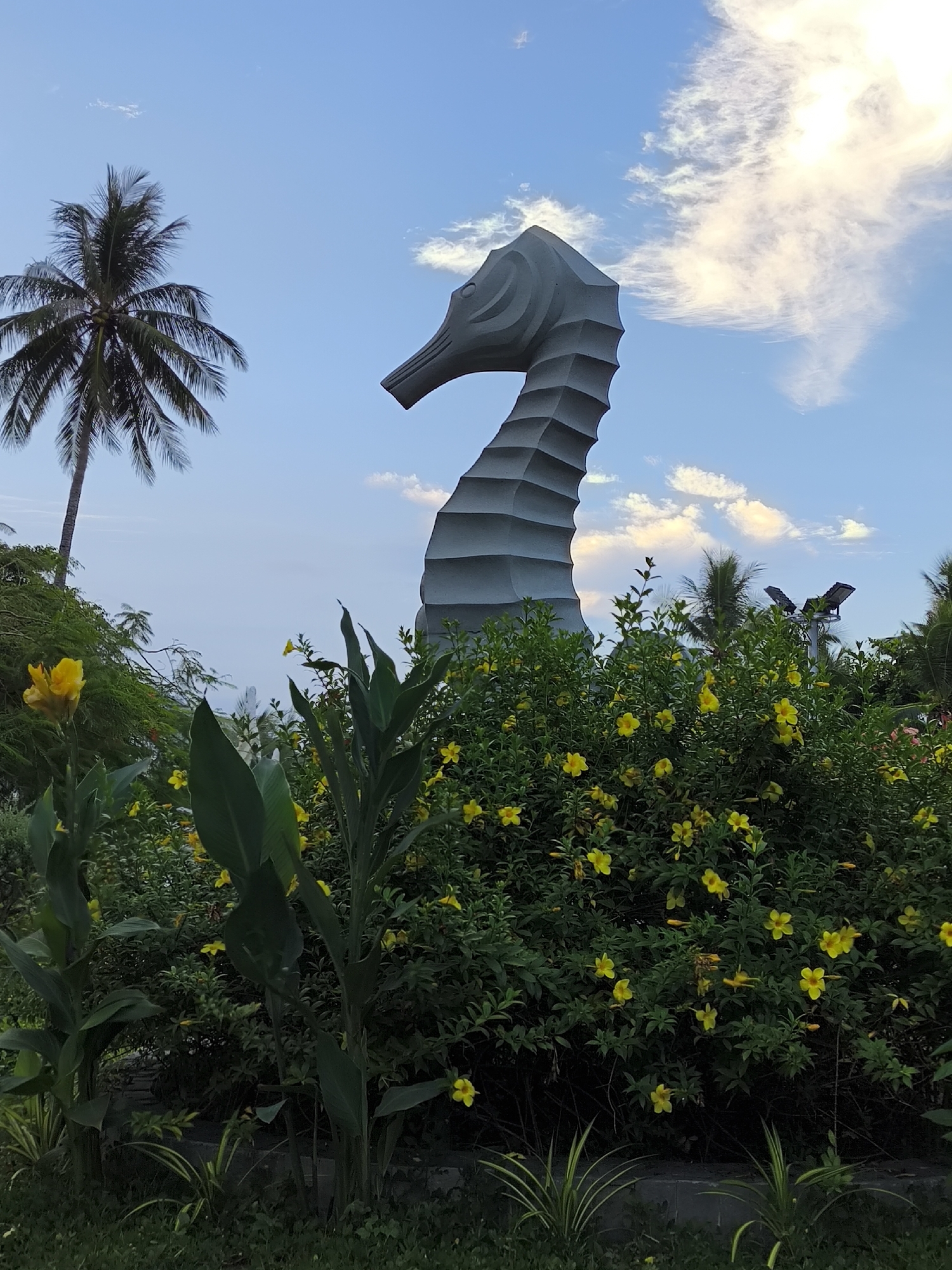 This media file depicts an outdoor scene featuring a large, stylized seahorse sculpture against a backdrop of a partly cloudy sky. The sculpture, made of a light-colored material, has a segmented, almost fin-like texture along its body and neck. In the foreground, lush green foliage and vibrant yellow flowers create a dense border. A tall palm tree stands to the left of the sculpture, its fronds reaching towards the sky. The lighting suggests it is either early morning or late afternoon, with the sun casting a warm glow on the clouds. The overall atmosphere is peaceful and serene, evoking a sense of tropical beauty. The location is identified as Bac Nha Trang Ward, Vietnam, which aligns with the presence of palm trees and tropical flora. There is no visible text in the image.