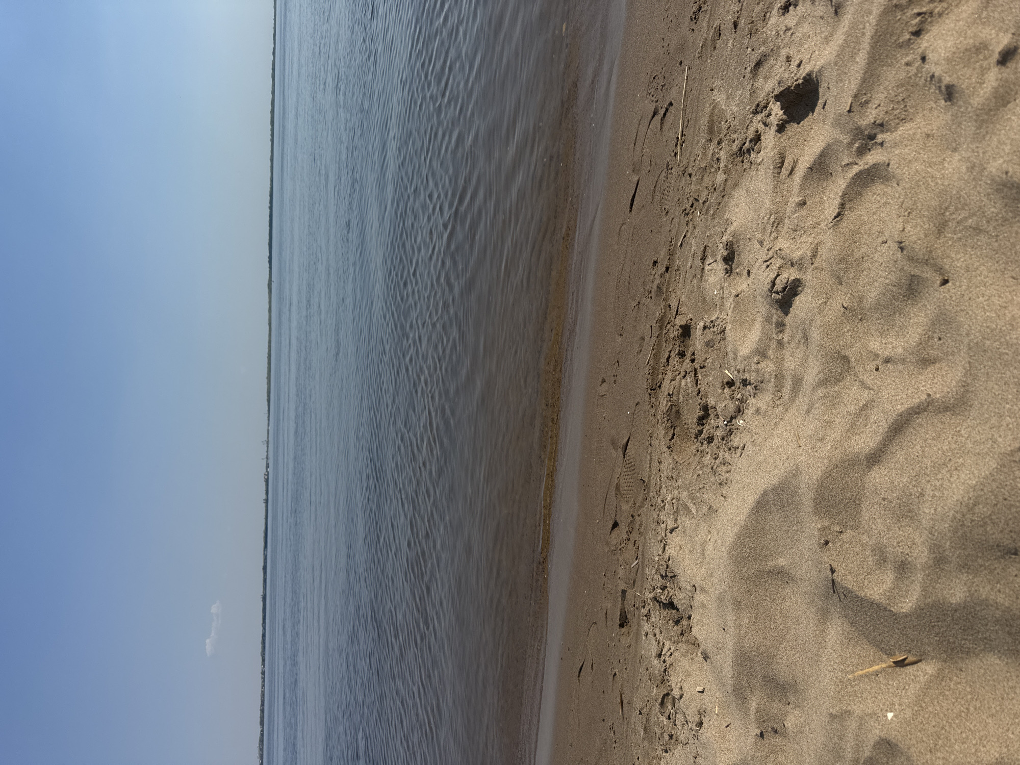 The image depicts a shoreline. To the left, water extends to the horizon. The sky is pale blue. To the right, sand covers the ground, with visible footprints and small pieces of debris. Given the prompt's information, the location is likely a beach near Saratov, Russia. No people are visible.