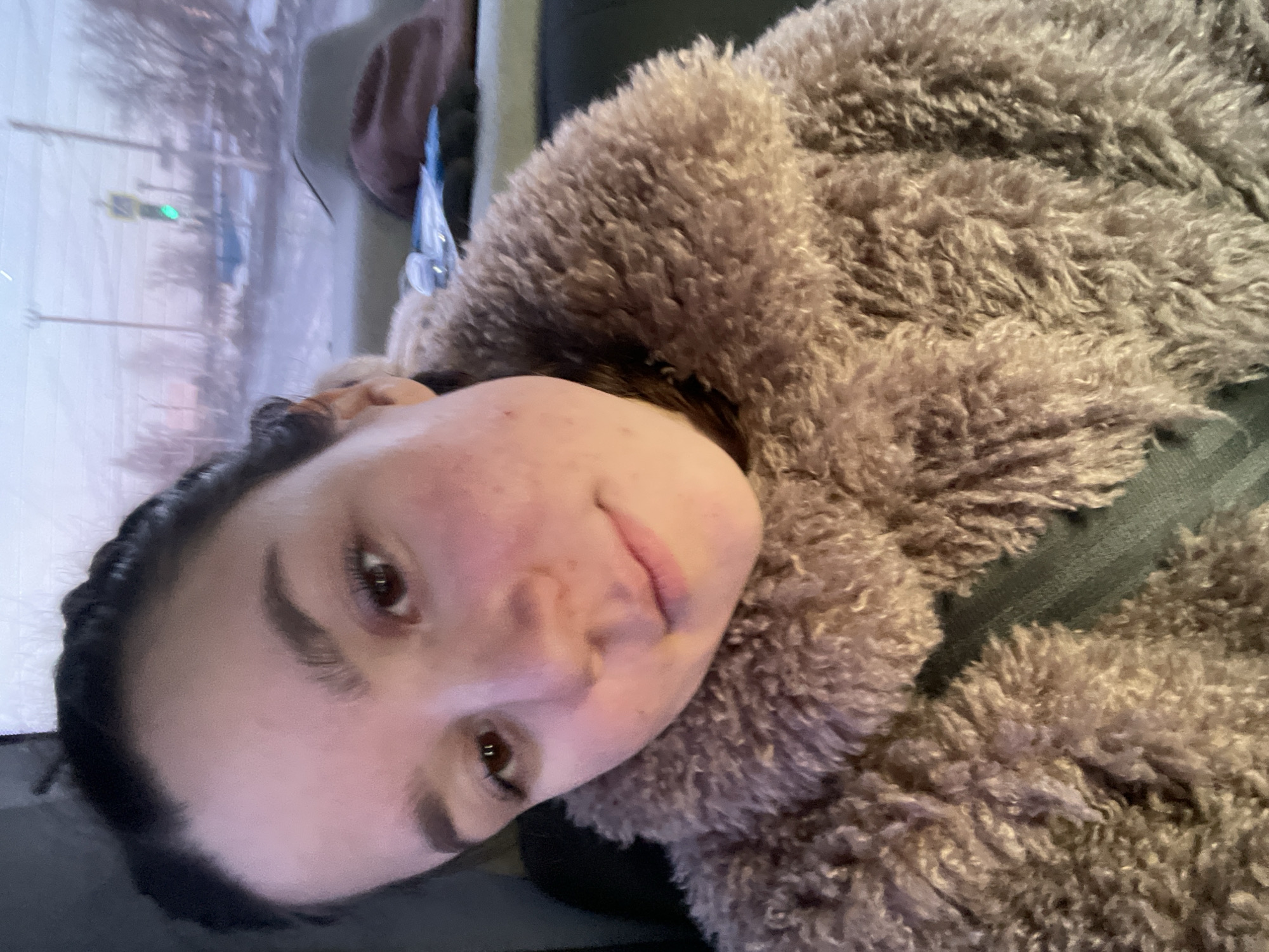 The image displays a close-up, chest-up view of a young female, approximately 18-25 years old, seated inside a vehicle. She has dark hair, dark brown eyes, and a fair complexion with minor facial blemishes on her cheeks and forehead. She is looking directly at the camera with a neutral expression. She is wearing a thick, textured, light brown or beige faux fur coat, with a dark seatbelt strap visible across her chest.
The interior of the vehicle shows dark gray or black upholstery behind and to the left of the subject. On the seat adjacent to her (to the viewer's left), a dark brown wide-brimmed hat and other indistinct objects, including a blue item and a shiny metallic object, are present.
Through the vehicle's window, a blurred winter street scene is visible, consistent with an urban or semi-urban area in Chaikovsky, Russia. The background features several utility poles, bare trees, and ground covered in snow or ice. A green traffic light signal is clearly discernible in the upper right portion of the background, along with a yellow square street sign. The overall lighting is dim, suggesting a winter day with overcast skies, or dawn/dusk conditions.