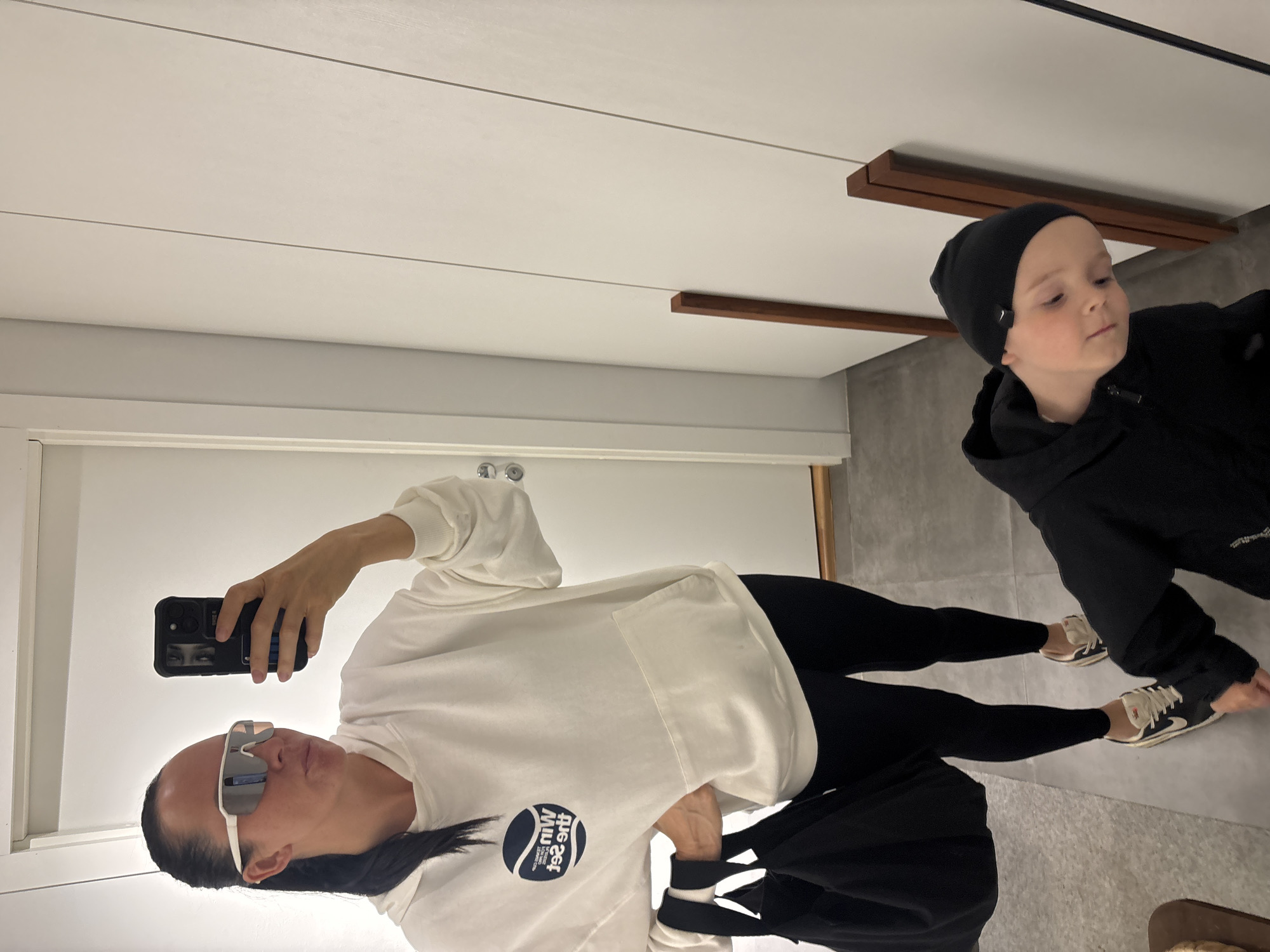A woman and a young child are in an indoor setting. The woman is holding a phone, taking a selfie. She is wearing sunglasses, a white sweatshirt with a logo, and black leggings. The child is wearing a black beanie and a black jacket. The room has a white ceiling with dark wood accents and gray flooring. A door is visible to the left. Based on the information available, the location is not identifiable as Chaikovsky, Russia.