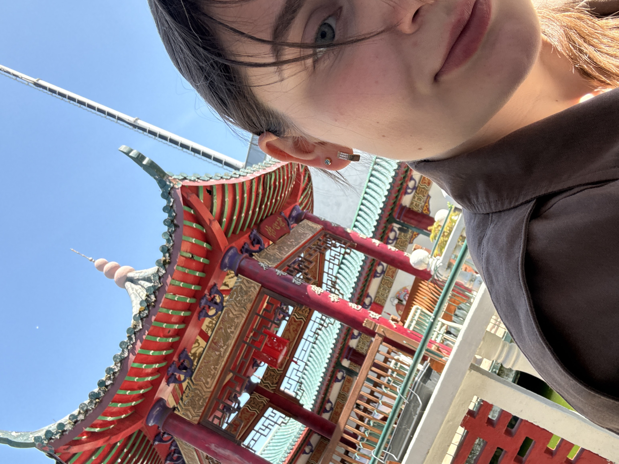 A close-up selfie captures a person in the foreground against the backdrop of a vibrant, traditional East Asian-style pagoda or pavilion. The person, a female, is visible on the upper right and top of the frame, showing her forehead, right eye, ear, nose, mouth, and chin. She has light skin, dark hair, and her right ear features an earring with a small blue stud and a longer, rectangular, possibly jeweled piece. She is wearing a dark, collared shirt.
The architectural structure dominates the lower and left-center of the image. It showcases a multi-tiered roof with distinctive red and green tiles, complemented by ornate red pillars. Golden panels adorned with intricate patterns and script-like characters are visible on the building's facade. Inside, a red lantern hangs amidst the detailed latticework. Decorative elements in shades of blue/purple and green add to the building's colorful aesthetic. The structure's spired top, featuring spherical ornaments, is visible against a clear blue sky. A tall, white pole or antenna extends into the sky on the upper left.
The scene is set outdoors under bright daylight, indicated by the clear blue sky and strong illumination. The overall impression is that of a travel photo or a visit to a culturally significant site. Given the context of Hainburg an der Donau, Austria, this building likely represents a specific landmark, such as a pagoda, situated within that European location.