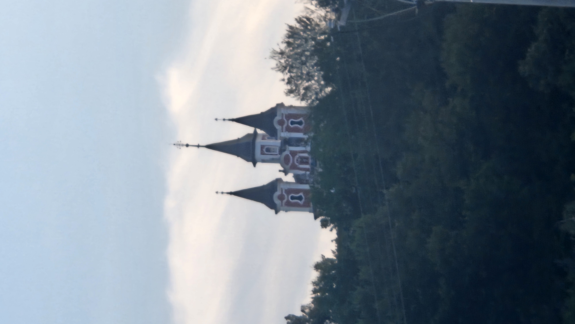 The image shows a church with three spires, situated on a hilltop surrounded by trees. The church has a distinctive architectural style with red and white facades and dark grey spires. The sky above is overcast with some visible clouds. The church is likely the Church of the Exaltation of the Holy Cross, also known as the Greek Catholic Cathedral, located in Prešov, Slovakia. The church is a prominent landmark in the city, and its elevated position on the hill makes it visible from many parts of Prešov. The image has a slightly hazy quality, suggesting it may have been taken from a distance or in low light conditions.