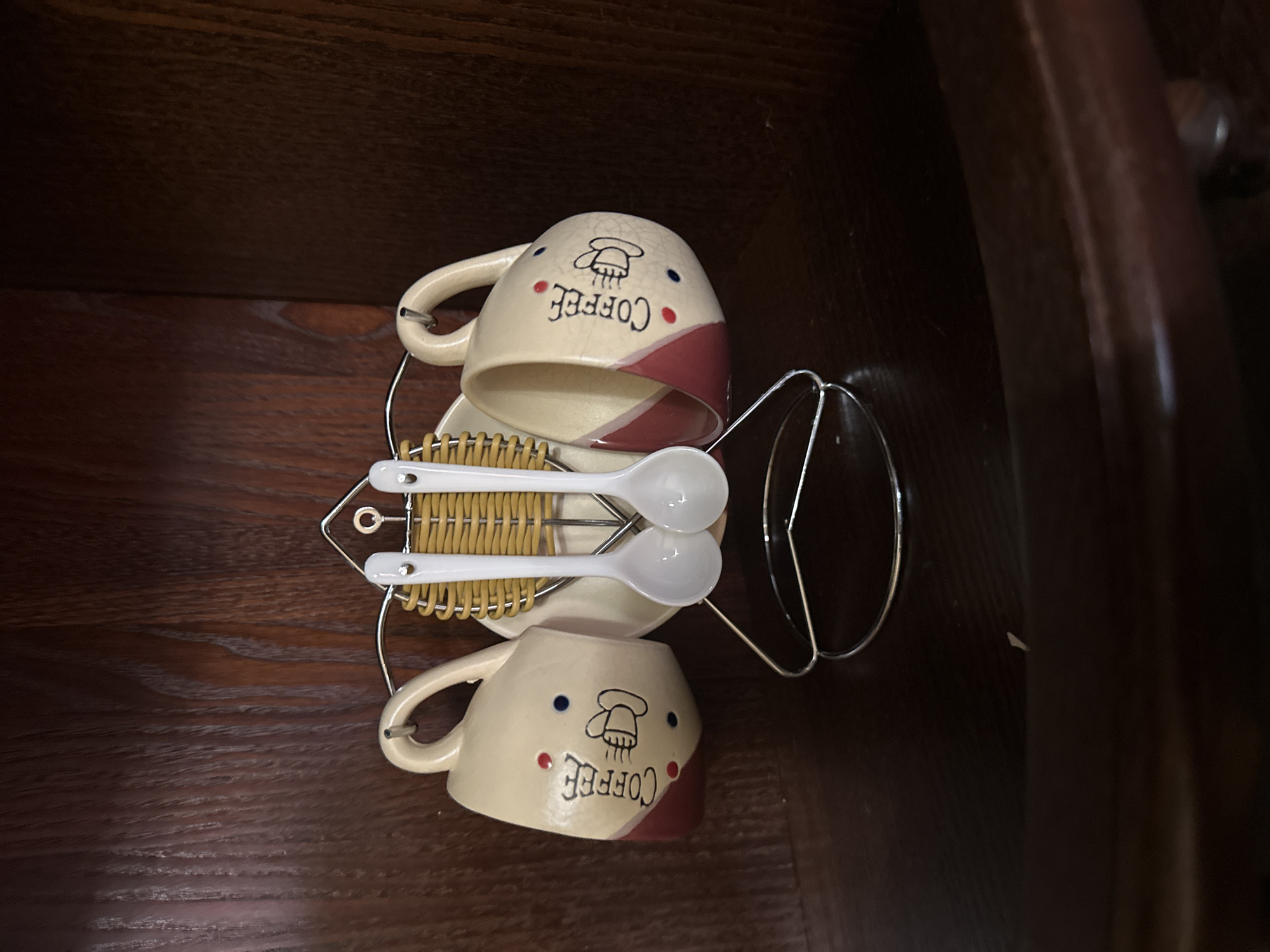 This is a close-up, eye-level, indoor shot of a coffee cup and spoon set displayed on a wooden shelf. The set includes two cream-colored mugs with red and blue polka dots and the word "Coffee" printed on them in black. Each mug has a handle and is adorned with a drawing of a steaming coffee cup. Two white spoons with yellow braided handles hang from a silver metal rack. The rack is supported by a circular base. The wooden shelf is dark brown with a visible grain pattern. The overall scene suggests a domestic setting, possibly in Aleksandrovka, Russia, where the items are being stored or displayed. There are no people or animals present in the image.