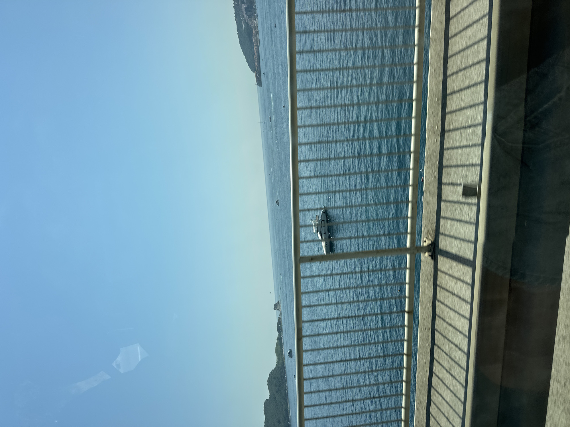 The image captures a view from a bridge overlooking the Adriatic Sea near Budva, Montenegro. The foreground features a metal railing and a concrete walkway, casting distinct shadows on the ground. Beyond the bridge, the turquoise water stretches to the horizon, dotted with several boats and yachts. In the distance, lush green hills rise from the coastline, with buildings and structures visible on the slopes, suggesting the presence of a town or city. The sky is clear and blue, indicating a sunny day. One prominent white yacht is visible in the middle ground, close to the railing. The overall scene depicts a serene coastal landscape under a bright, clear sky.