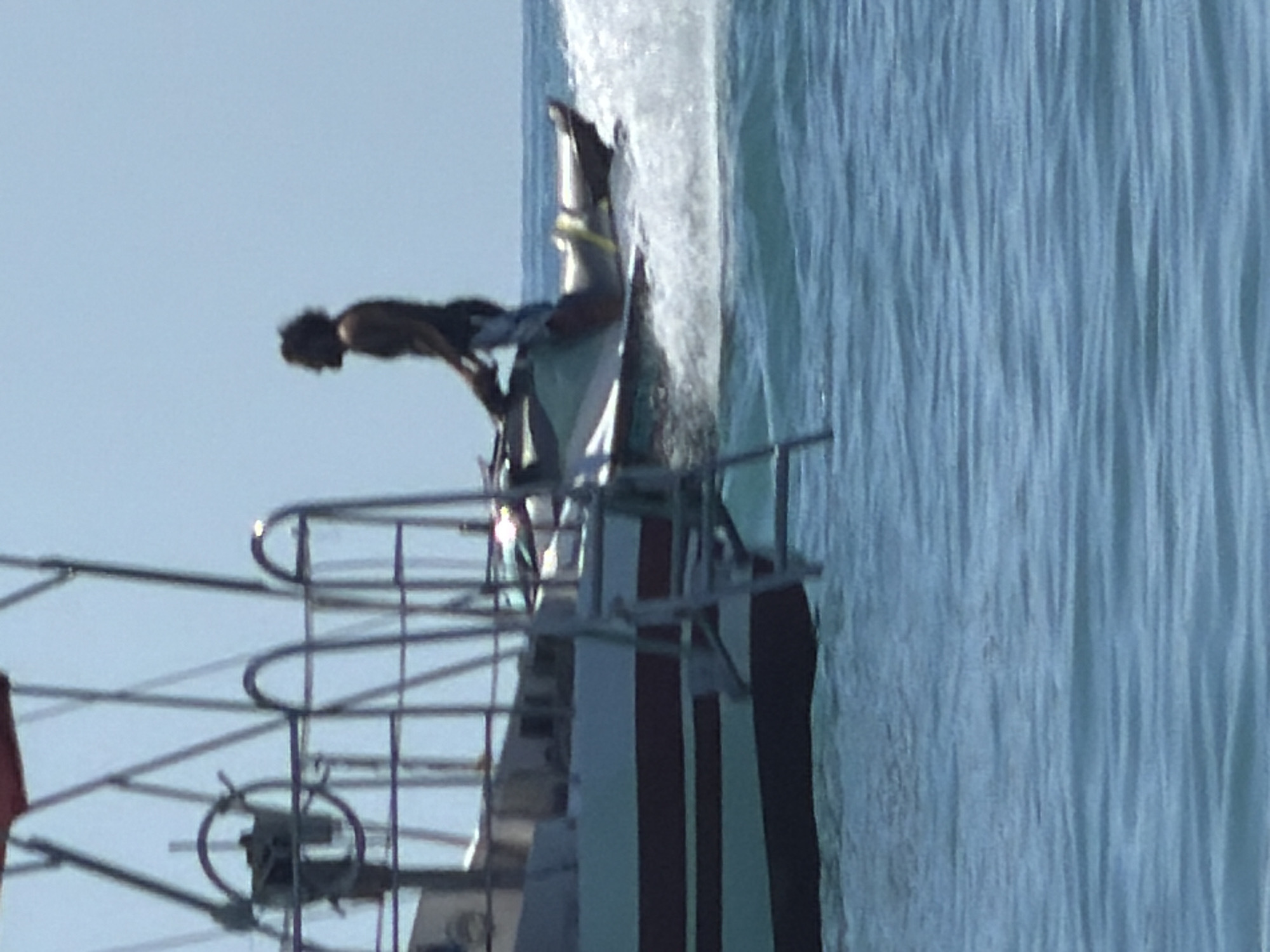 A man is riding a jet ski in the ocean, creating a large splash of water. He is wearing dark shorts and is shirtless, with dark curly hair. He is leaning forward, holding onto the handlebars of the jet ski. The jet ski is white with some green accents. In the background, a portion of a boat is visible, with a red canopy and a ladder attached to its side. The boat appears to be anchored or moving slowly. The water is a bright blue, and the sky is a clear, light blue. The scene appears to be set on a sunny day. There is no information available to suggest this image was taken in Sochi, Russia.