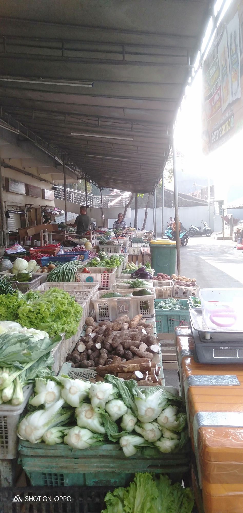 This is a medium shot, taken from a low angle, of an outdoor market stall in South Jakarta, Indonesia. The market is covered by a corrugated metal roof supported by metal poles. Fluorescent lights are attached to the underside of the roof. In the foreground, there are several crates of fresh vegetables, including lettuce, bok choy, and what appear to be taro roots and cassava. To the right, a digital scale is placed on top of stacks of orange crates. In the midground, two men are standing at their respective stalls, one in a dark green t-shirt and the other in a striped t-shirt. They appear to be vendors, interacting with customers or arranging produce. Behind them, more produce is displayed in crates, and further back, a green trash bin is visible. Beyond the market stalls, a street is visible with parked motorcycles, one of which is blue and being ridden by two people. The background shows a white wall and some trees. The overall impression is a bustling, open-air market scene on a sunny day.