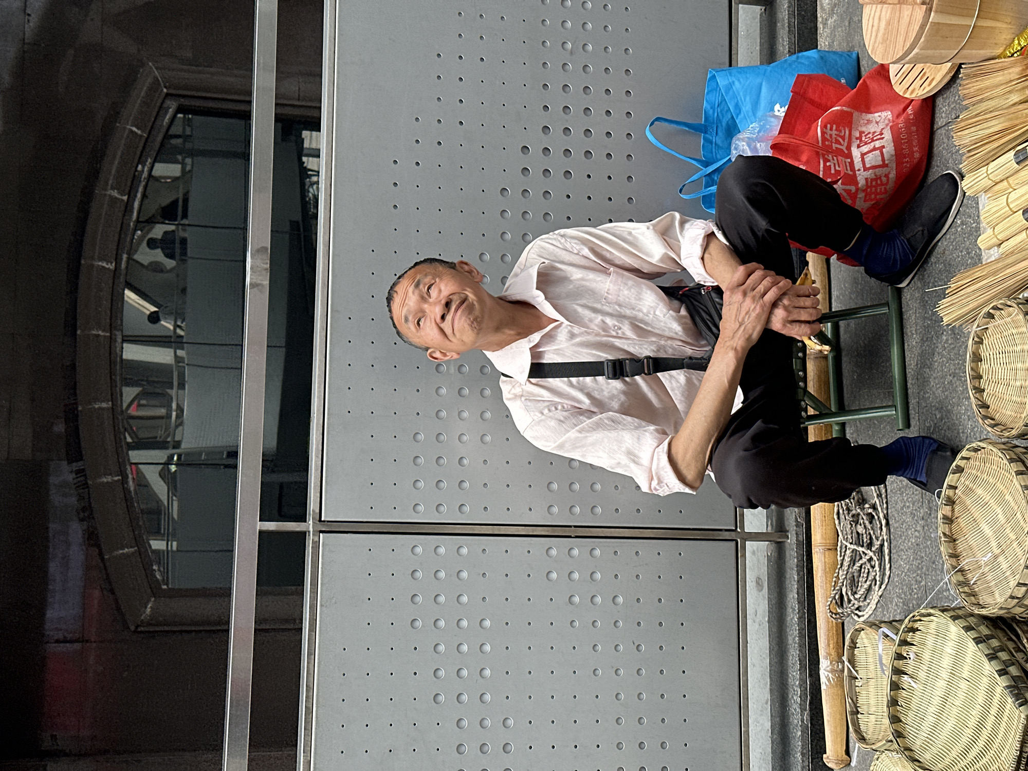 A horizontal, ground-level shot depicts a man sitting outside in Jiulongpo District, China. The man is middle-aged, wearing a light pink button-down shirt, black pants, black shoes, and blue socks. He wears a black bag across his torso. The man sits on a green metal folding chair. He is turned to face the viewer with a slight smile.
To the right of the man are red and blue tote bags, wooden objects, and several woven baskets. To the left of the man is a gray metal wall with an array of small, circular holes. A window with a reflective surface is located to the far left of the frame.