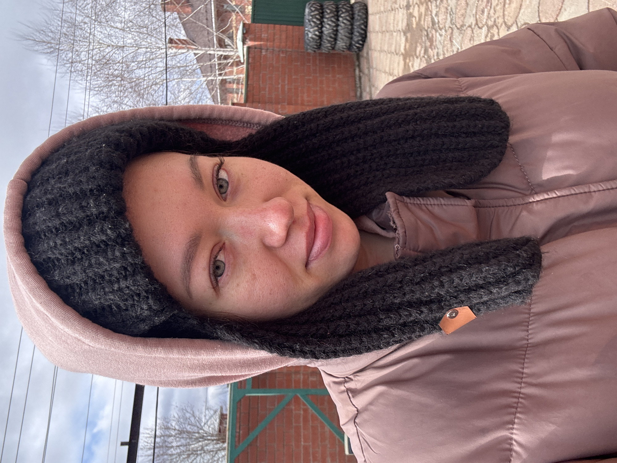 A person is wearing a mauve puffer jacket with a brown hood lined with a dark fuzzy material. The person is positioned close to the camera, focusing on their face. They have light skin, green eyes, and a neutral expression with a slight smile. Behind the person, a brick building with green accents is partially visible. The sky appears overcast with bare trees visible in the background. A stack of tires can be seen behind the building. The image was taken in a sideways orientation, with the top of the frame showing sky and the bottom showing part of the jacket. The location is Ферма, Russia.