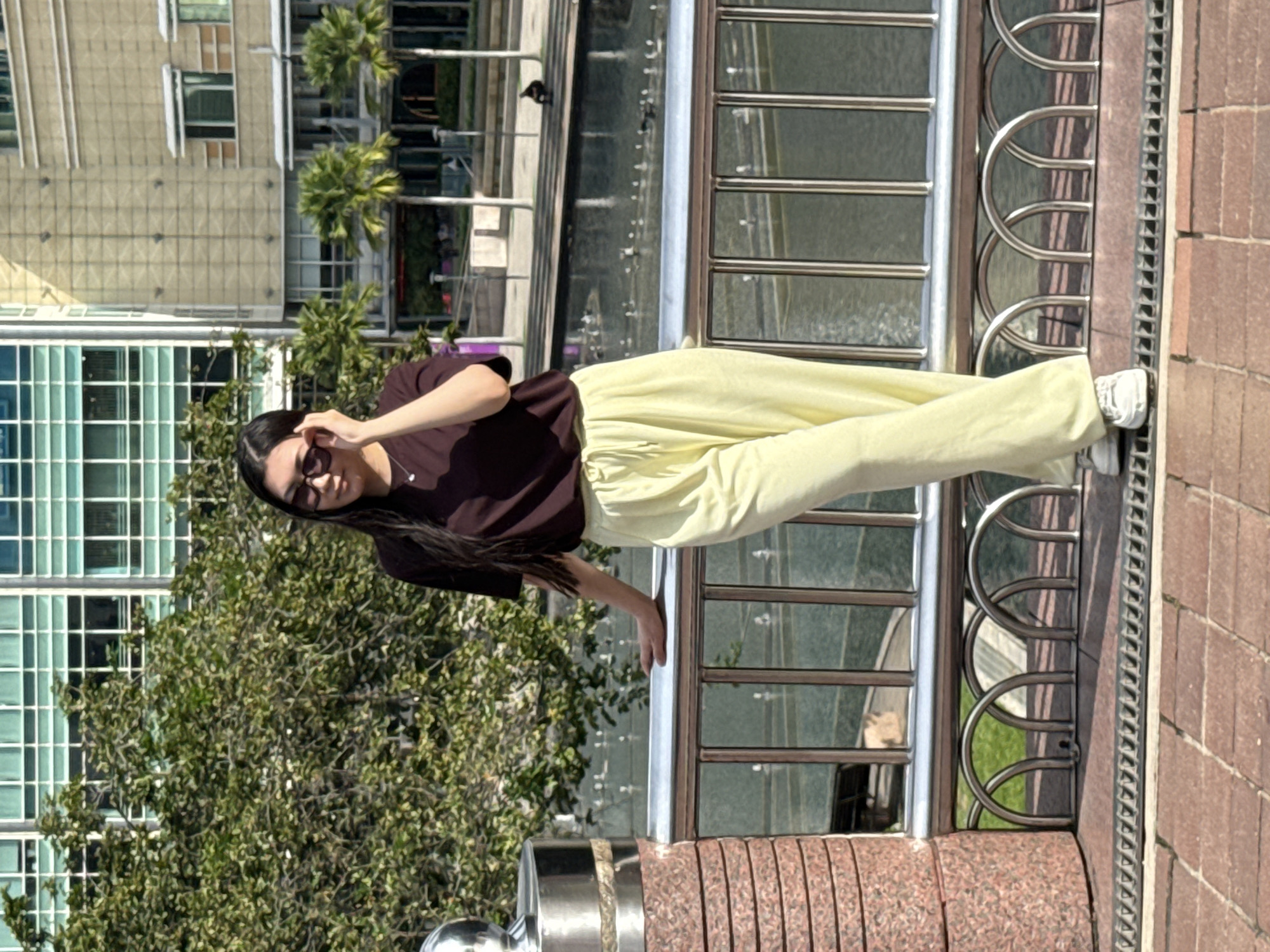 A young woman wearing sunglasses, a brown t-shirt, and light yellow trousers is posing next to a metal railing. She is standing outdoors on a brick-paved surface. Behind the railing, a body of water reflects the buildings and sky, suggesting a canal or river. In the background, there are modern buildings, including one with a grid-like facade, and palm trees. Based on the architectural style and presence of palm trees, the location is likely Kuala Lumpur, Malaysia.