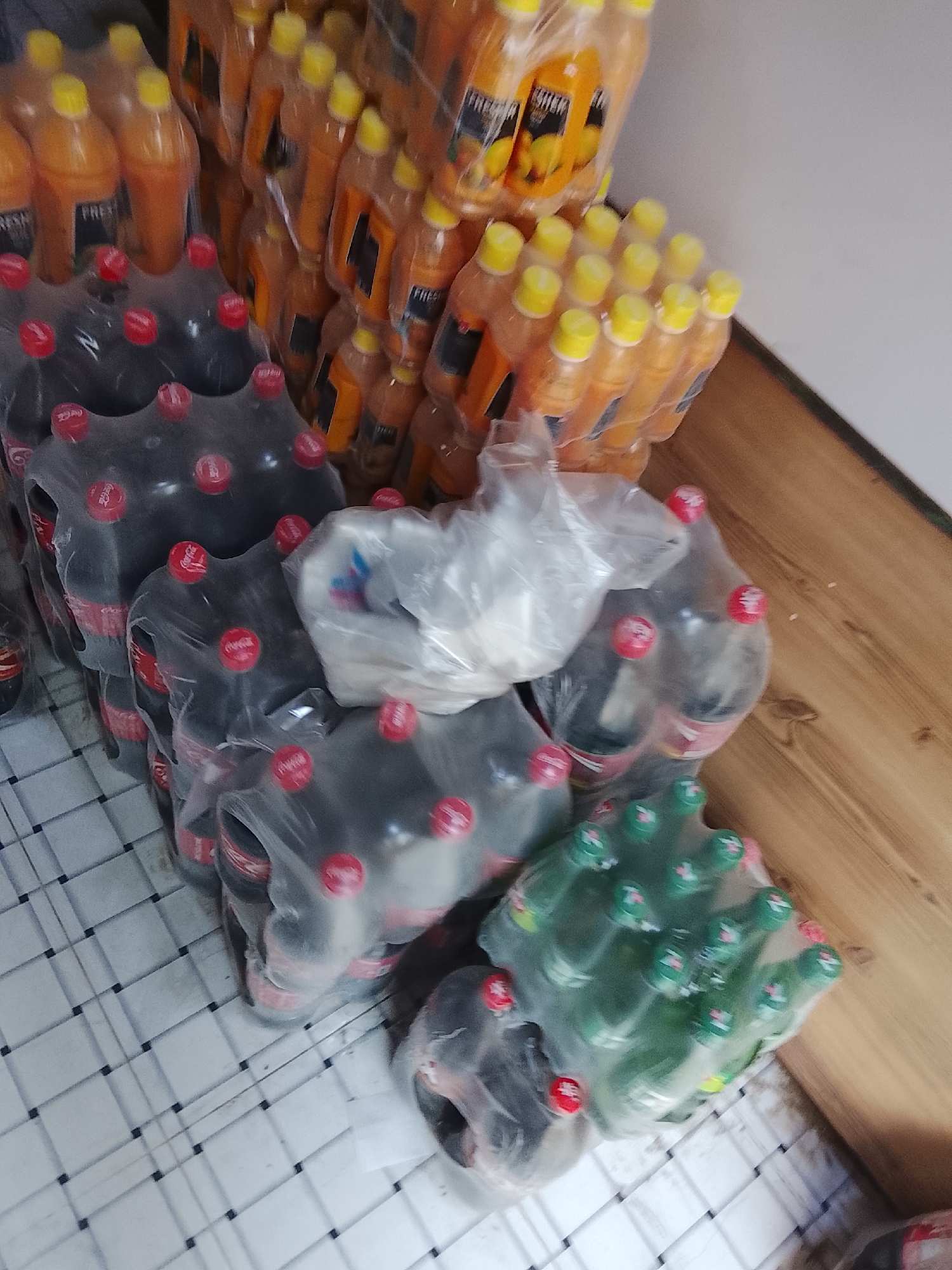 The image depicts stacks of shrink-wrapped beverages on a floor. There are stacks of orange juice bottles, dark soda bottles (likely Coca-Cola), clear soda bottles, and green soda bottles, all appearing new and stacked for storage or sale. The location may be in Gulgasht Colony, Pakistan. The floor is a checkered pattern, and there's a wooden surface along the edge of the frame.