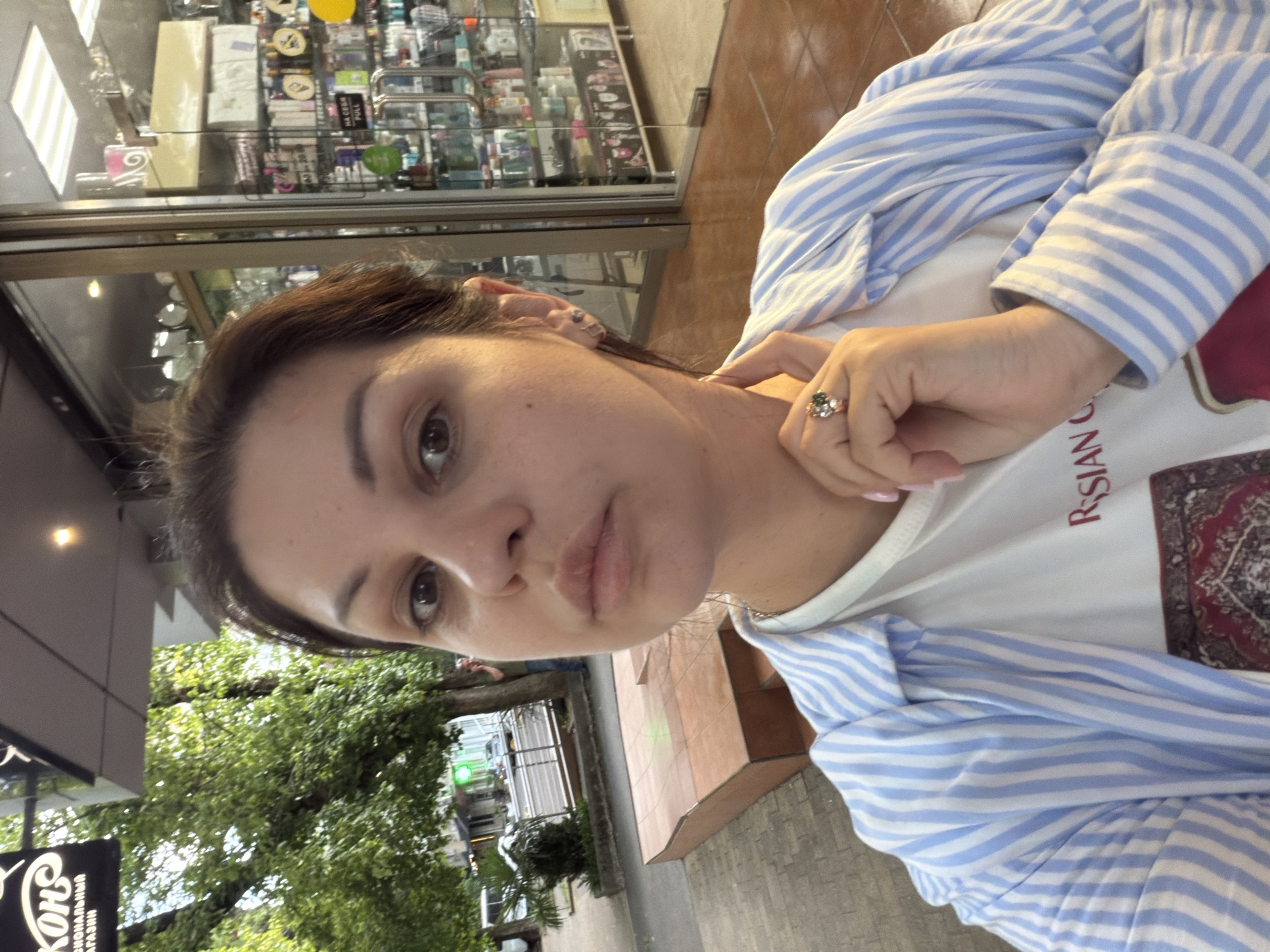 A medium shot captures a woman with brown hair and brown eyes taking a selfie outdoors. She is wearing a blue and white striped shirt over a white t-shirt with red text and a patterned design. A ring with green and clear stones is visible on her right hand, which is touching her neck.
The background suggests an urban environment, possibly a commercial street. To the left, trees and buildings are visible, along with a sidewalk and a street with vehicles. To the right, the entrance to a store or pharmacy is apparent, with glass doors and shelves stocked with various products displayed inside. The flooring in front of the store appears to be tiled. The overall lighting suggests it is daytime, with some overcast conditions. The location appears to be a public area in Sochi, Russia, given the context of the request.