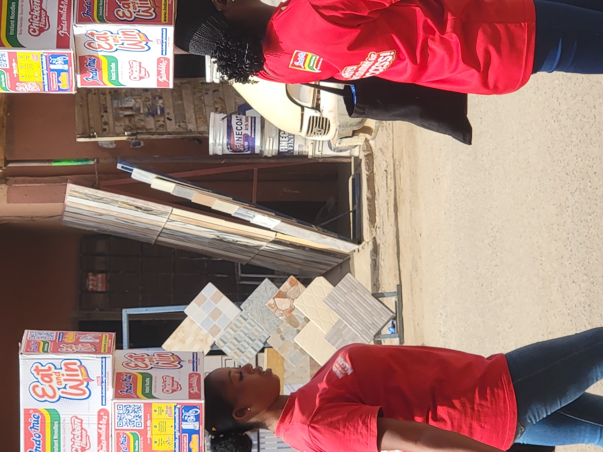 This outdoor image captures a commercial scene, likely a market stall or roadside shop, in Maiburiji, Nigeria, during daylight hours. The bright sunlight suggests a clear, sunny day.
Two individuals, possibly women, are visible. The person in the foreground wears a red t-shirt and blue jeans, with dark hair tied back. Another person in the background also wears a red top, potentially an apron, over a dark outfit, and has dark, curly hair. Both individuals appear to be facing away from the camera, engaging with the display.
Prominently featured are multiple stacked boxes of "Indomie Instant Noodles," displaying the phrases "Eat and Win" and "Chicken Flavor." Some boxes also show a QR code. The person in the background has a logo that reads "NASMI" and text that appears to be "Key For SUCCESS!" on their red top.
Behind the individuals, a diverse selection of ceramic floor and wall tiles is displayed vertically and horizontally. Further into the background, several paint buckets, labeled "FINECOAT" and "FINE," are visible. A white scooter or motorcycle is partially seen behind the person in the background. The ground appears to be a mix of concrete and dirt, and a brownish building wall forms the backdrop. The setup suggests a promotional event or a retail point for various goods, including noodles, tiles, and paint.