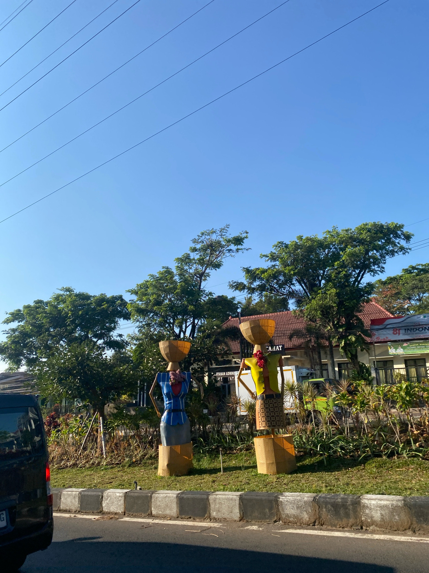 Two large straw figures, designed to resemble women carrying baskets on their heads, stand on a grassy median in front of a building. The figure on the left is adorned with a blue and white striped top and a grey skirt, while the figure on the right wears a yellow top and a patterned skirt. Both figures have straw baskets balanced on their heads. Behind the figures, a building with a tiled roof and signs is visible. The scene is set against a clear blue sky with power lines crisscrossing the upper portion of the image. A black van is parked to the left, and other vehicles and trees are present in the background. The figures appear to be decorative installations along a roadside.