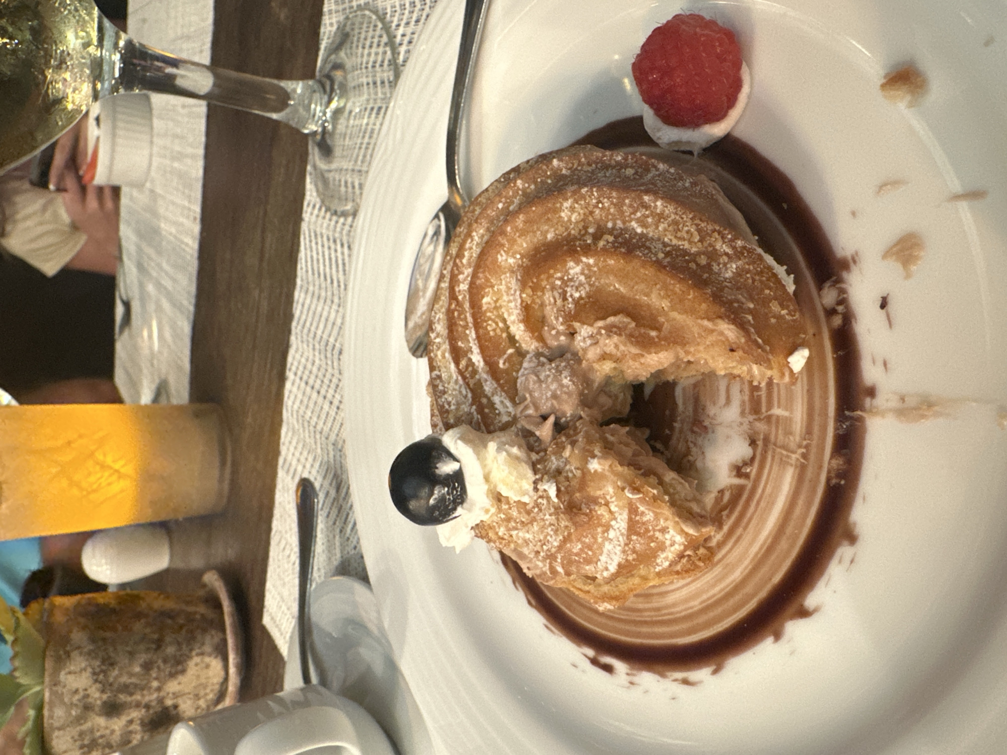A partially eaten pastry, likely a profiterole or similar cream-filled puff pastry, sits on a white plate with a swirl of dark chocolate sauce. The pastry is topped with a dollop of white cream, a single raspberry, and a single black fruit (possibly a blackberry or dark cherry). A silver spoon rests on the plate to the left of the pastry.
The scene appears to be set on a table in a restaurant or dining area. Part of a glass of light-colored liquid, possibly wine or a cocktail, is visible in the upper left, along with a glimpse of a person's hand. Other table settings, including a small pot, a butter knife, and possibly a salt shaker, are also present. The tablecloth has a light-colored, textured pattern.
Without additional information, it is not possible to confirm the precise location in Playa del Carmen, Mexico, or to provide specific details about the people involved.