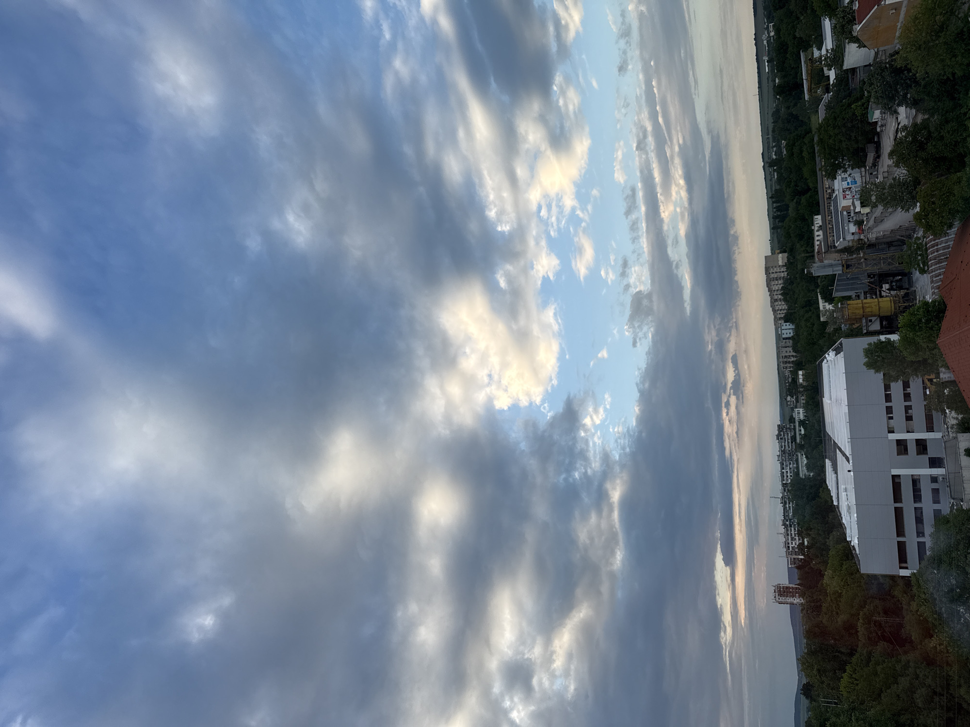 The image depicts a cityscape under a cloudy sky, with a clear patch of blue visible. Predominantly gray and white clouds occupy the majority of the frame. Buildings are visible in the background, interspersed with trees. In the lower right corner, a large white building is partially visible. Further away, there are residential buildings and construction cranes, indicating urban development. The location is identified as Lovech, Bulgaria, based on the availability of contextual information. The overall setting suggests a late afternoon or early evening, given the lighting conditions. No people are visible in the image.